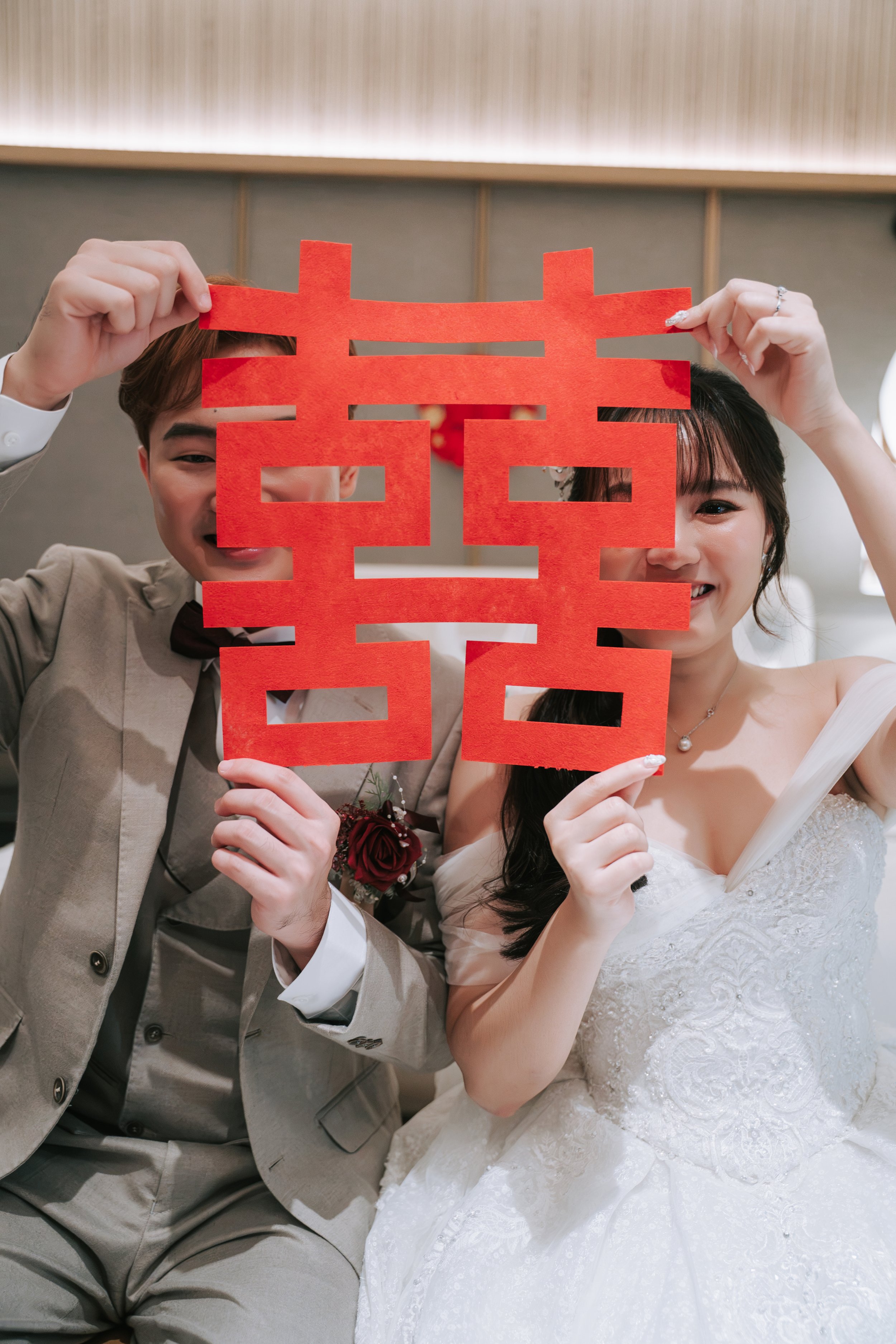 A bride and groom holding a red decorative paper cutout in front of their faces at a wedding celebration.