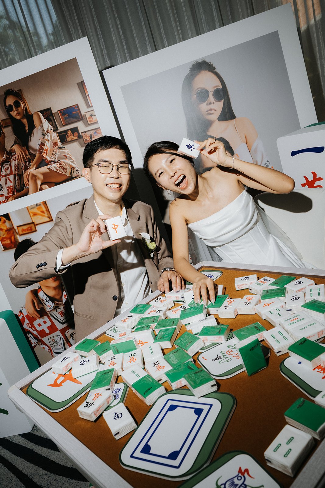 A newlywed couple celebrating their wedding with a card game at a table filled with Mahjong tiles and cards. The groom is wearing a beige suit, glasses, and a boutonnière, while the bride is in a white dress holding a card near her head. They are smiling and making playful gestures, surrounded by large photographs of women wearing sunglasses on the backdrop.