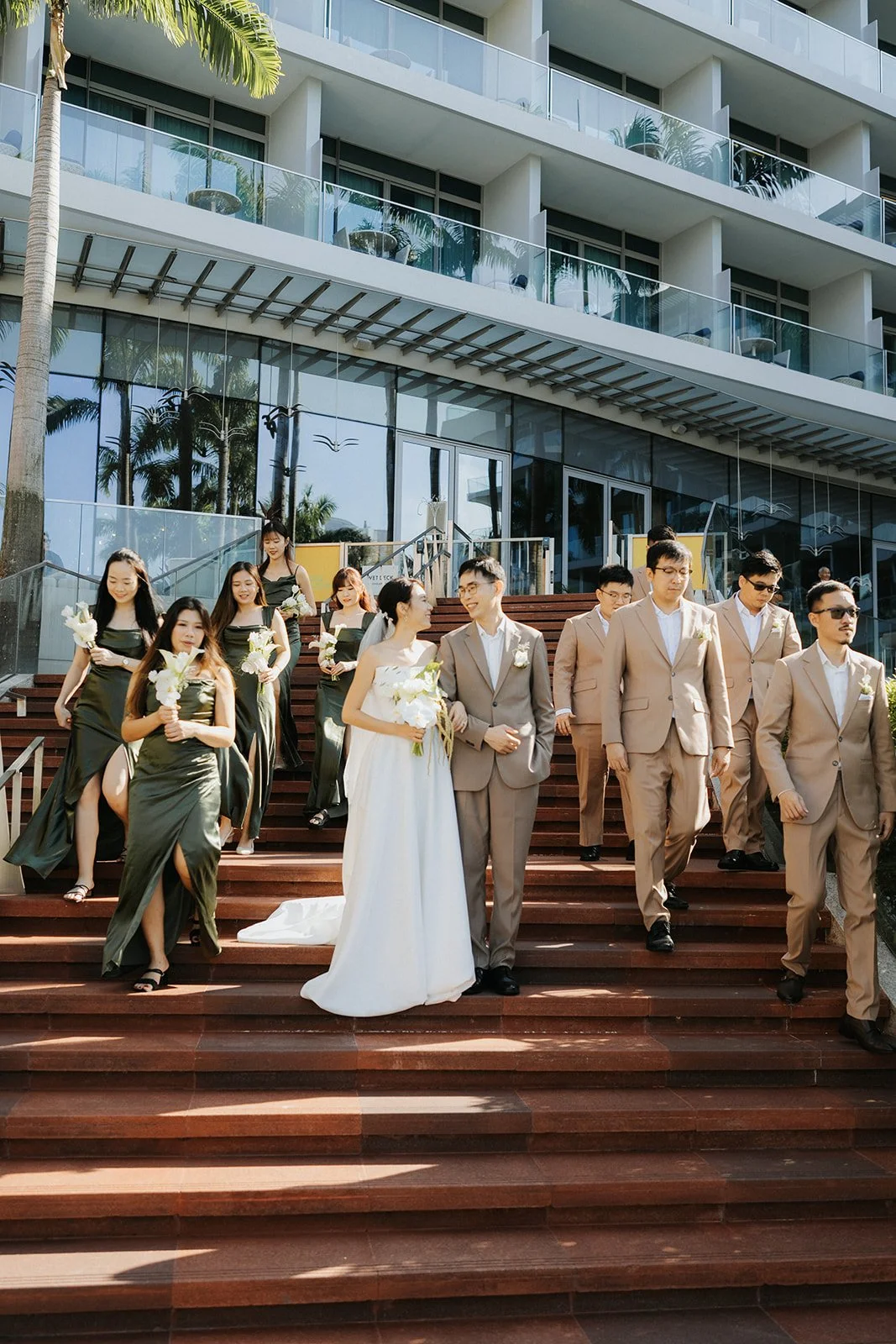 A group of people in wedding attire walking down steps outside a modern building with glass balconies, palm trees, and blue sky.