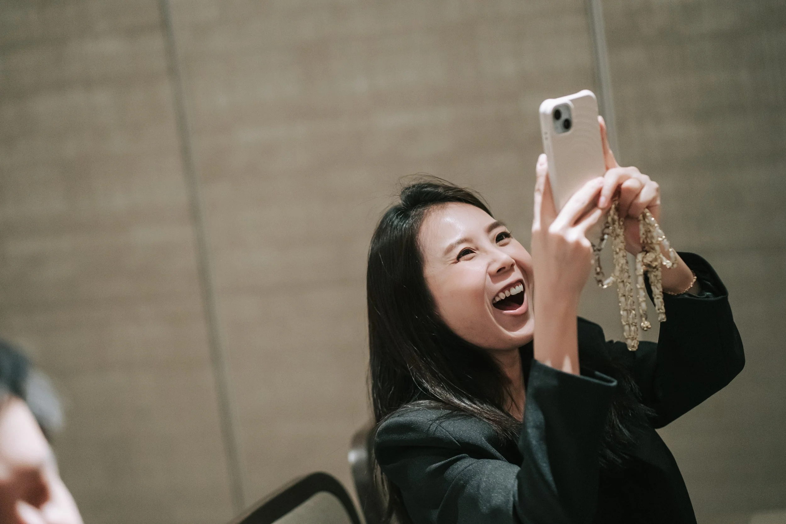 A woman is smiling and taking a selfie with her phone, holding it with her right hand, in an indoor setting with a gray wall in the background.