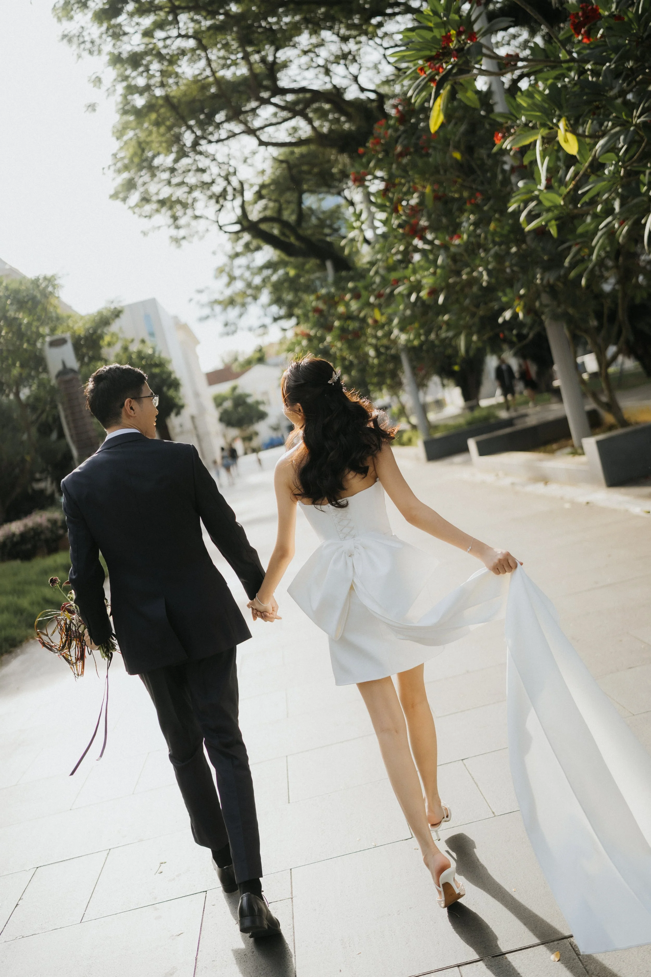 A bride and groom walking hand in hand outdoors, with trees and a building in the background. The bride wears a white wedding dress with a full skirt and high heels, while the groom is dressed in a black suit and glasses, holding a bouquet of flowers in his left hand.