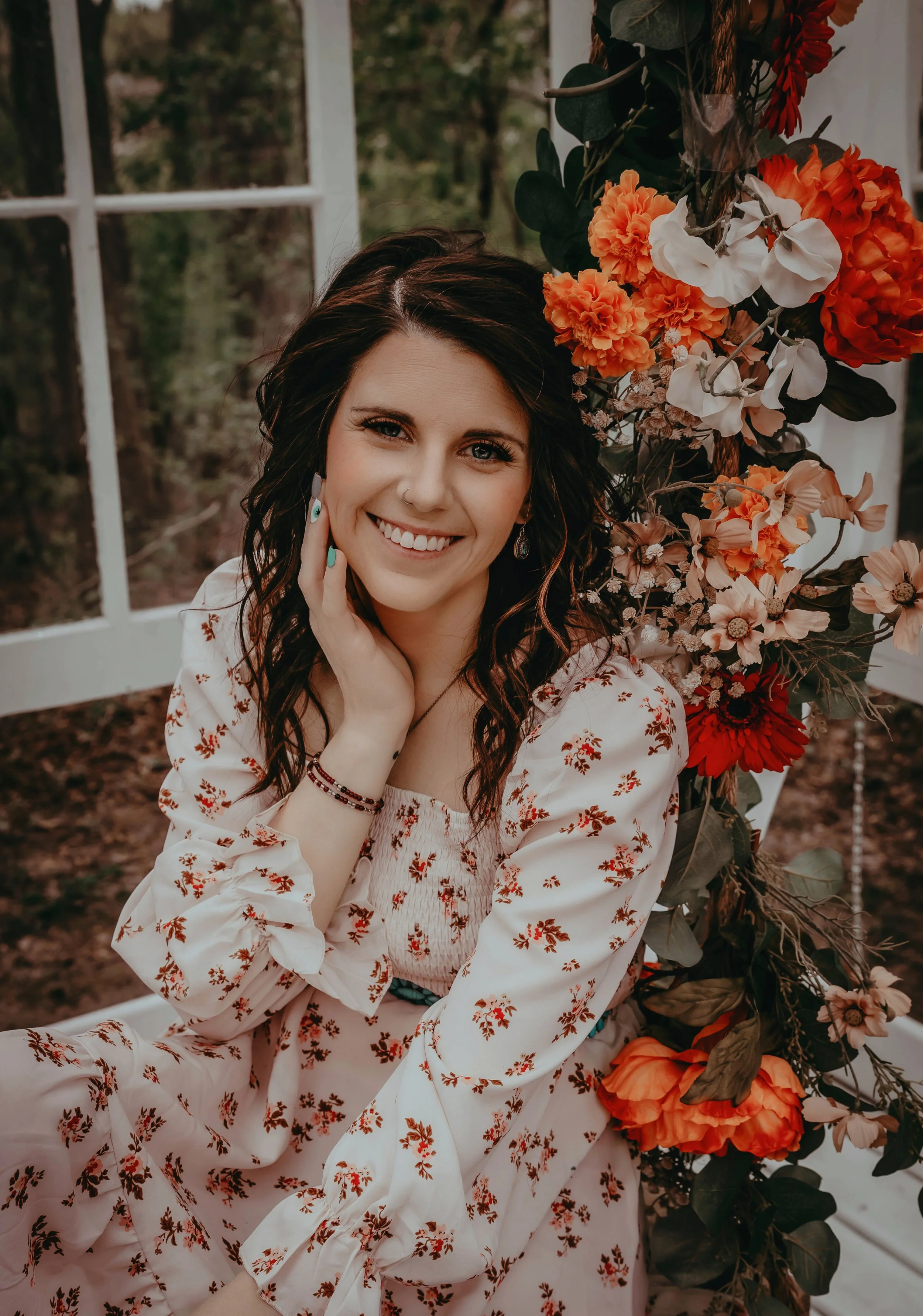 A woman with dark wavy hair, wearing a white floral dress with red flowers, sitting next to a bouquet of orange, red, and white flowers, indoors near a large window with greenery outside.