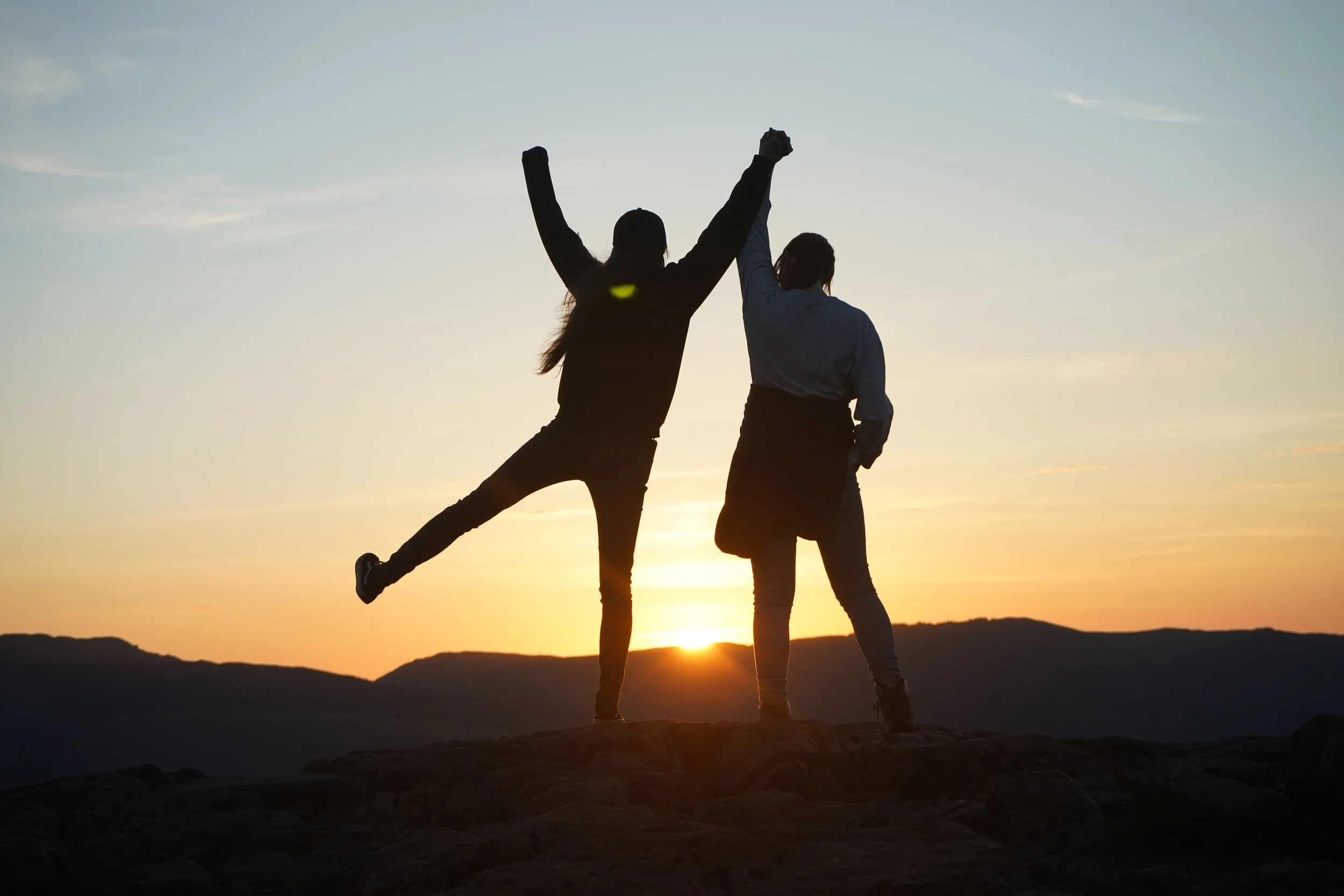 Two people outdoors looking at the sun poking up over a silhouetted hill.  One person has both their hands raised up holding one hand of the other person.