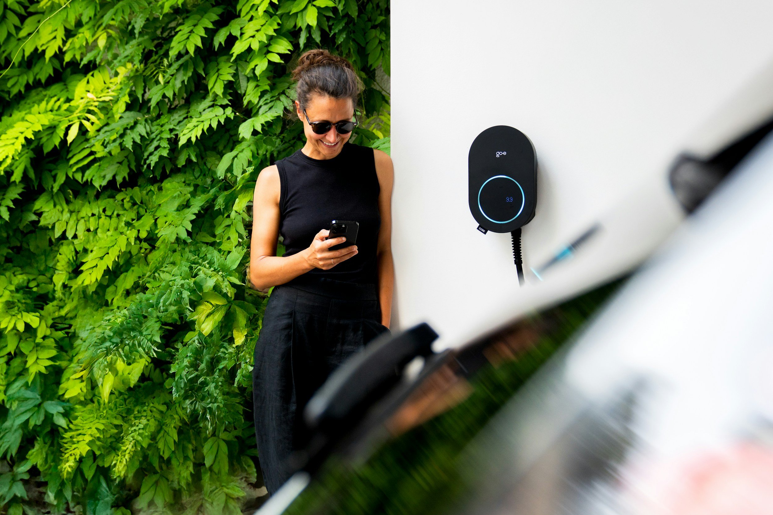 Une femme portant des lunettes de soleil, souriante, utilisant son téléphone, se tenant à côté d'une station de charge électrique pour voiture, avec un mur blanc et une abondance de feuillage vert en arrière-plan, vue partielle d'un appareil photo ou d'un microscope au premier plan.