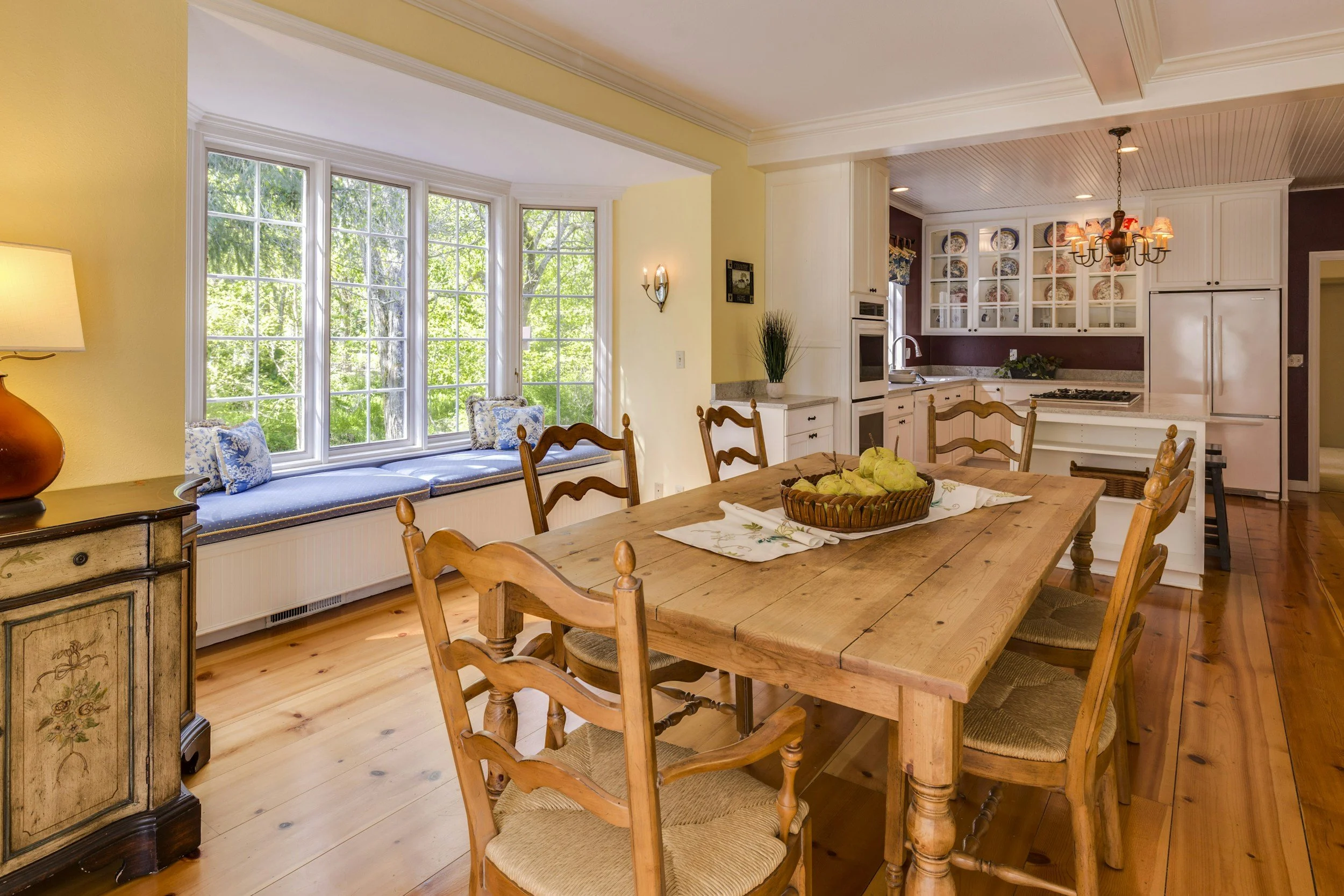 A cozy kitchen and dining area with a large window seat under multiple window panes, a wooden dining table with six chairs, and a kitchen with white cabinets and a chandelier.