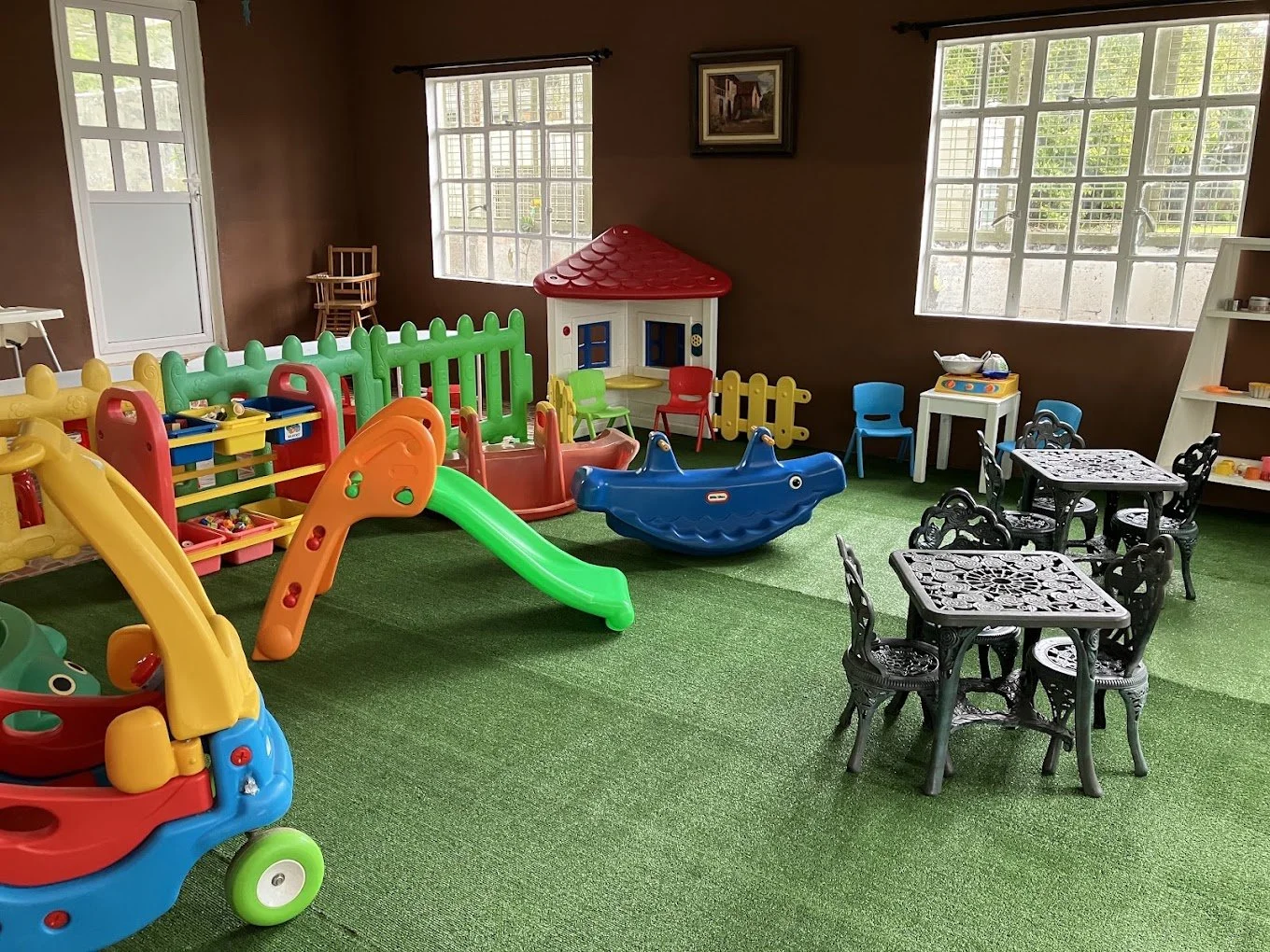 Salle de jeux pour enfants avec balançoires, toboggans, petites tables et chaises, et un tapis vert imitation herbe.