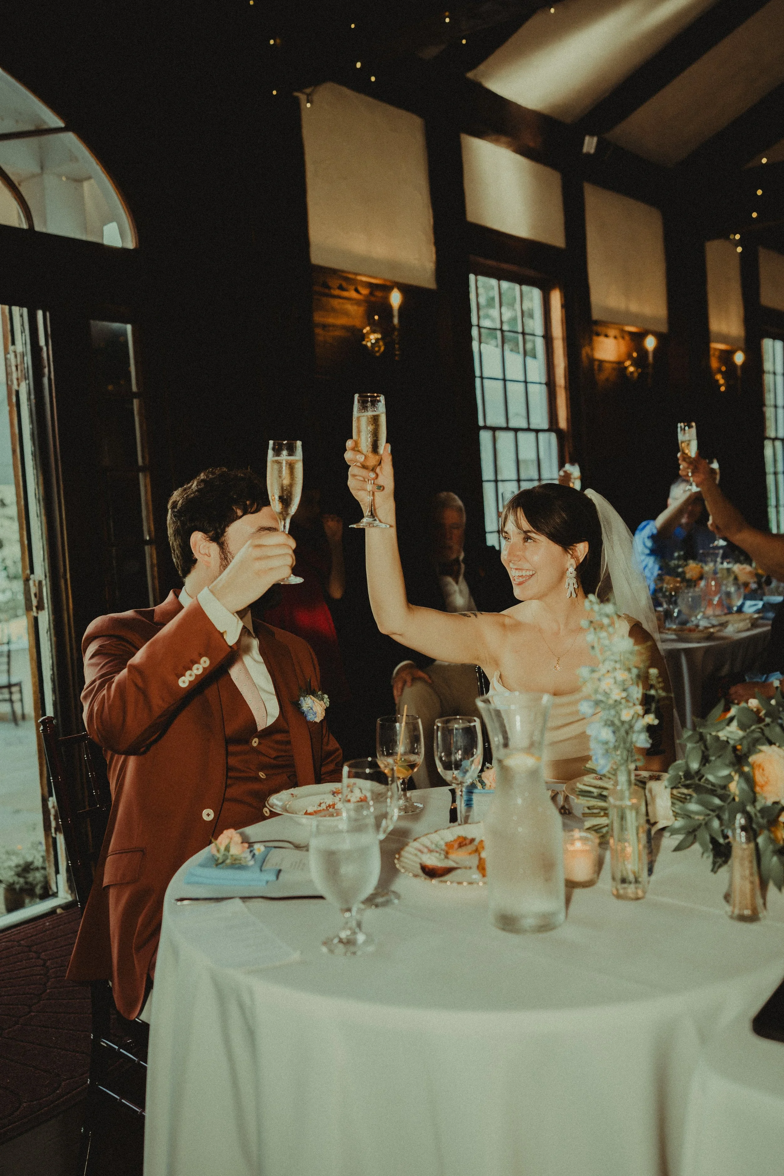 A bride and groom raising champagne glasses during a wedding toast at a celebratory dinner in a warmly lit venue with large windows and floral centerpieces on the table.