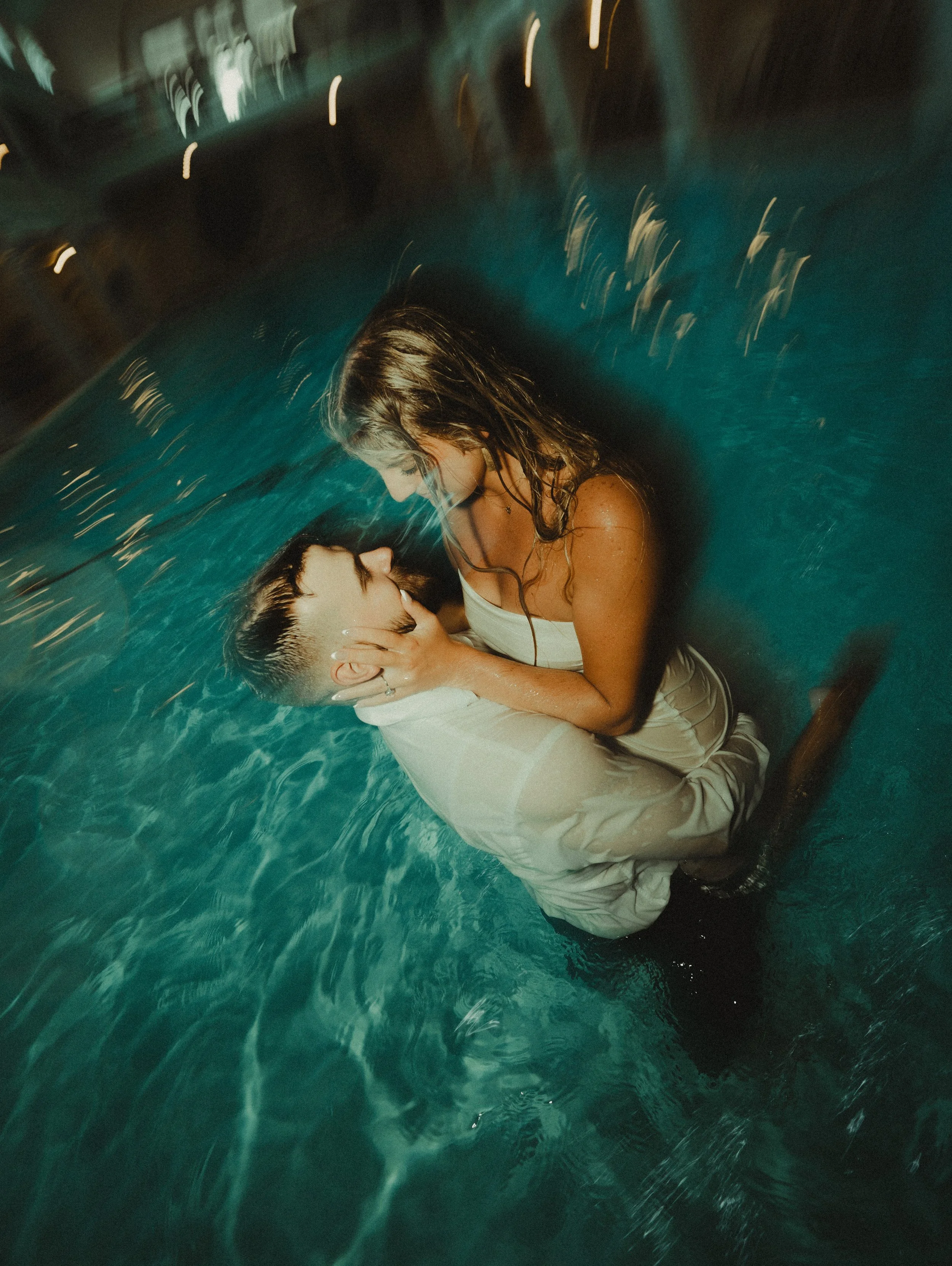 A woman in a white dress and a young boy are in a swimming pool, passionately kissing while holding each other's faces at night.