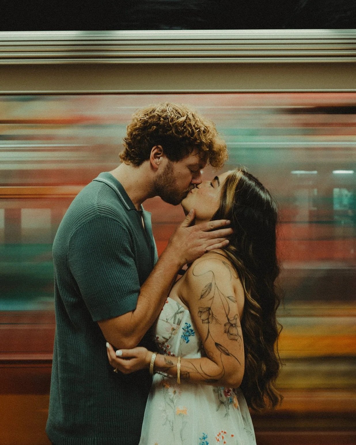 A young couple sharing a kiss in front of a moving train at a subway station, with vibrant colors and blurred background.