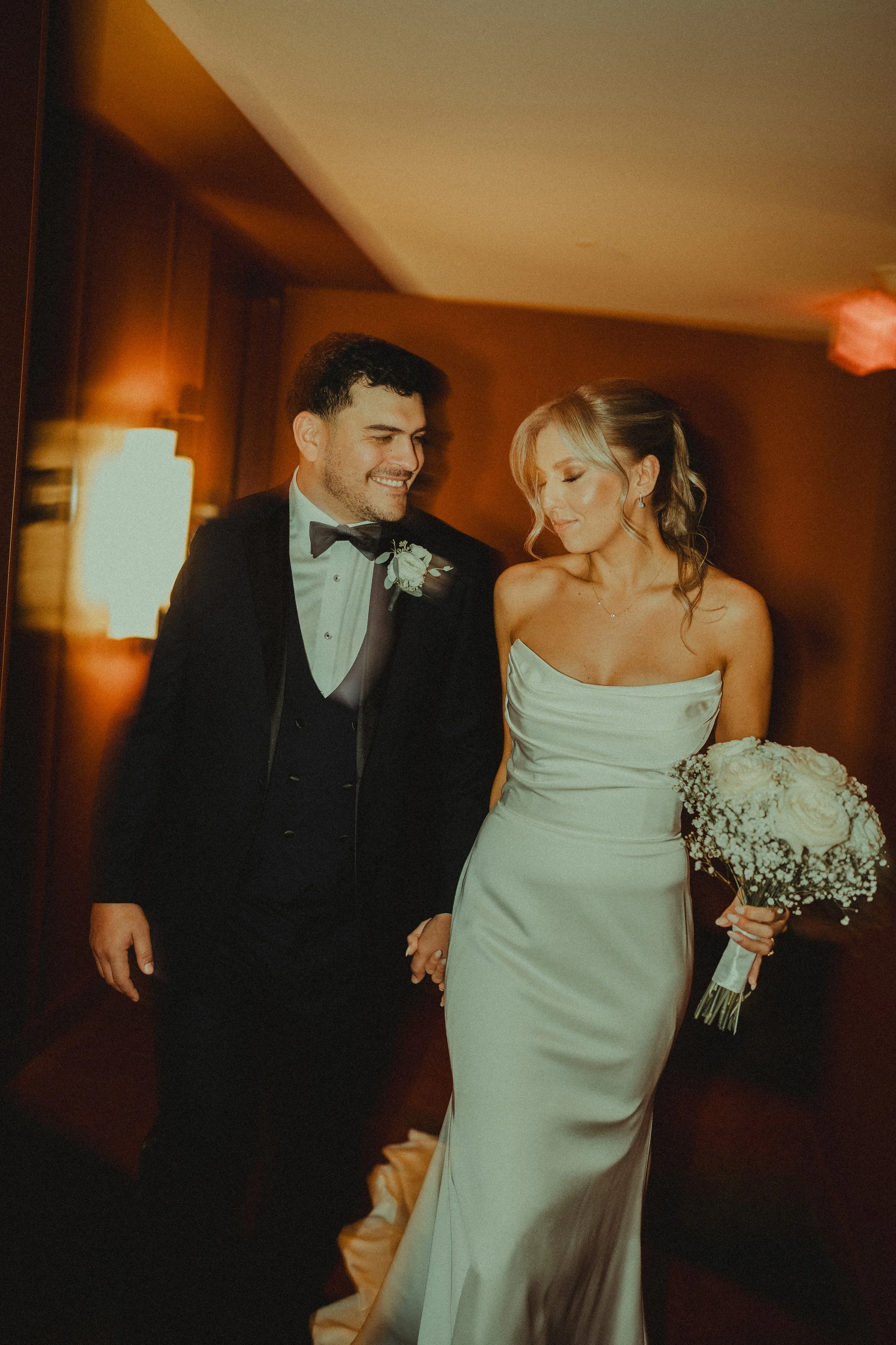 A smiling groom and a bride holding a bouquet, walking together at a wedding reception in an indoor setting.