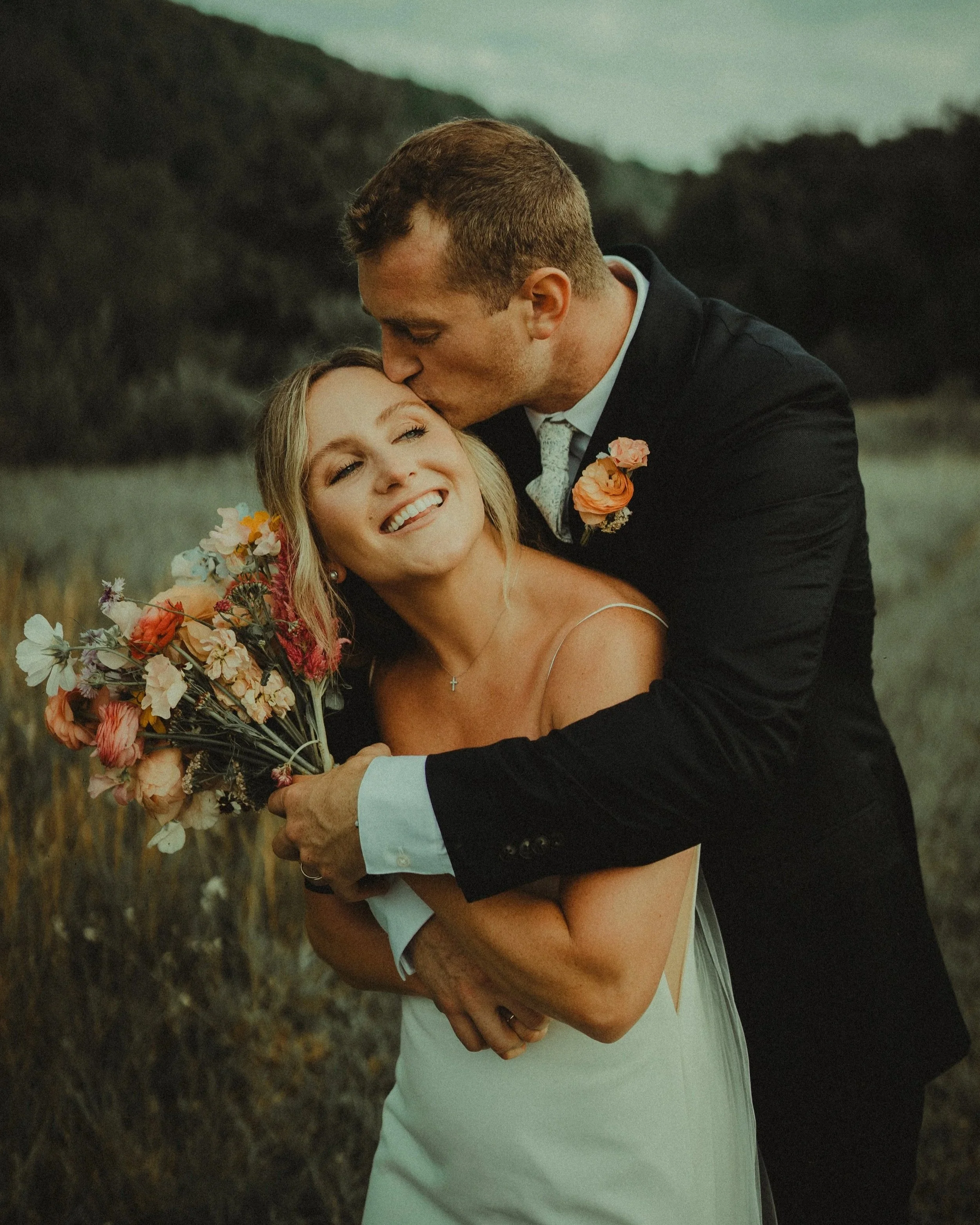 A couple, dressed in wedding attire, sharing an intimate moment outdoors. The man kisses the woman's forehead while she smiles. The woman holds a bouquet of flowers, and they are surrounded by a natural landscape with trees and mountains in the background.