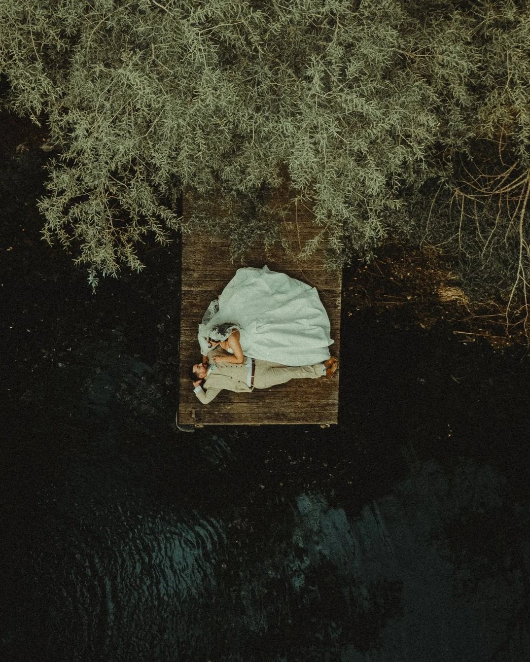 A couple lying on a wooden dock floating on water, surrounded by greenery.