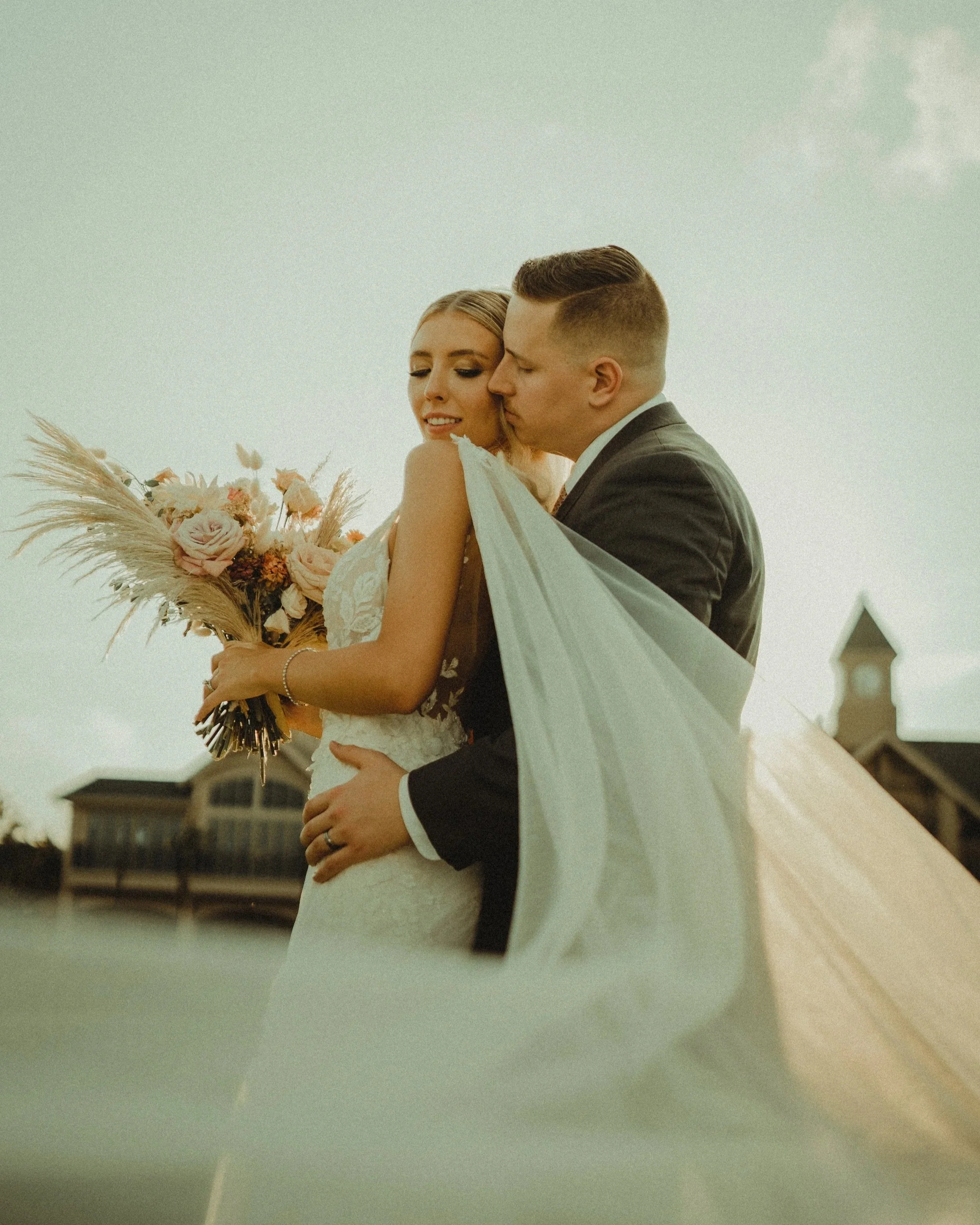 A newly married couple standing outdoors, with the officiant, possibly a wedding photographer, capturing a moment during their wedding ceremony.