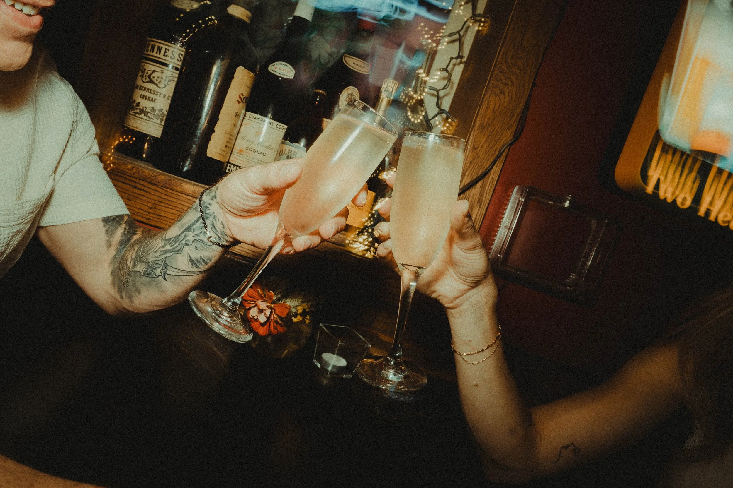 Two people clinking champagne glasses filled with a light-colored beverage in a bar or restaurant setting. A display of liquor bottles and a mirror are visible in the background, along with decorative string lights.