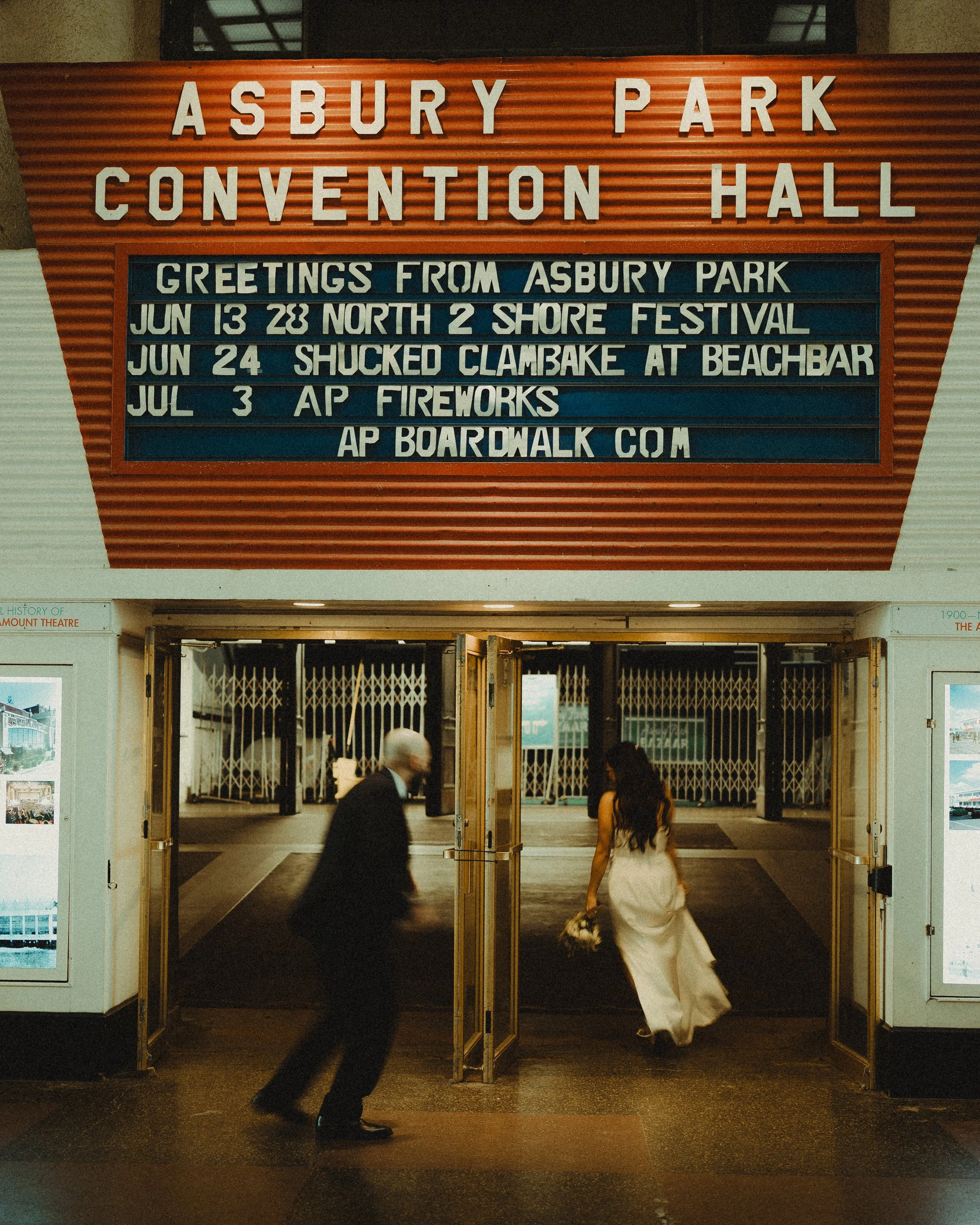 People entering the Asbury Park Convention Hall through open doors, with a sign above showcasing upcoming events and greetings from Asbury Park.