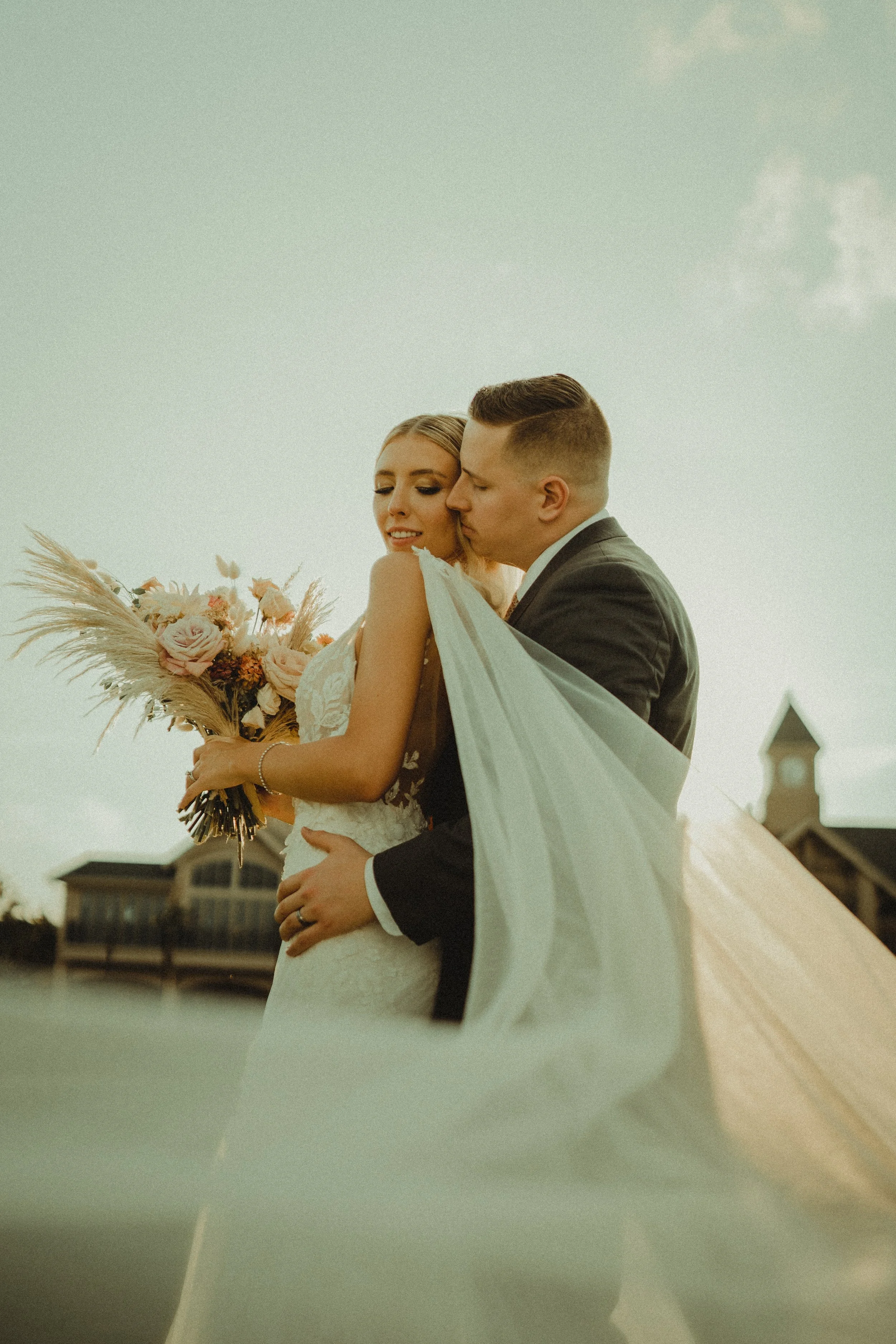 A bride and groom sharing a kiss during their wedding, with the bride holding a bouquet of flowers, and they are outdoors near a body of water with buildings in the background.