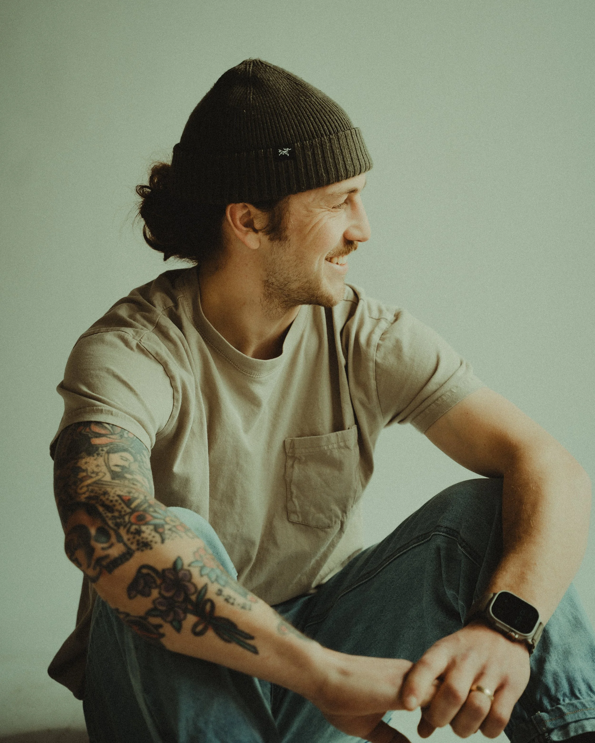 A man wearing a dark beanie, beige t-shirt, and black jeans, smiling, with tattooed arms, looking to the right, sitting against a plain background.
