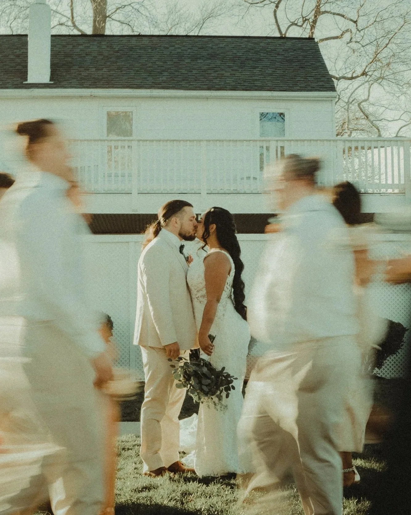 A couple in wedding attire sharing a kiss outdoors, surrounded by blurred people in white clothing walking past, with a white house and trees in the background.