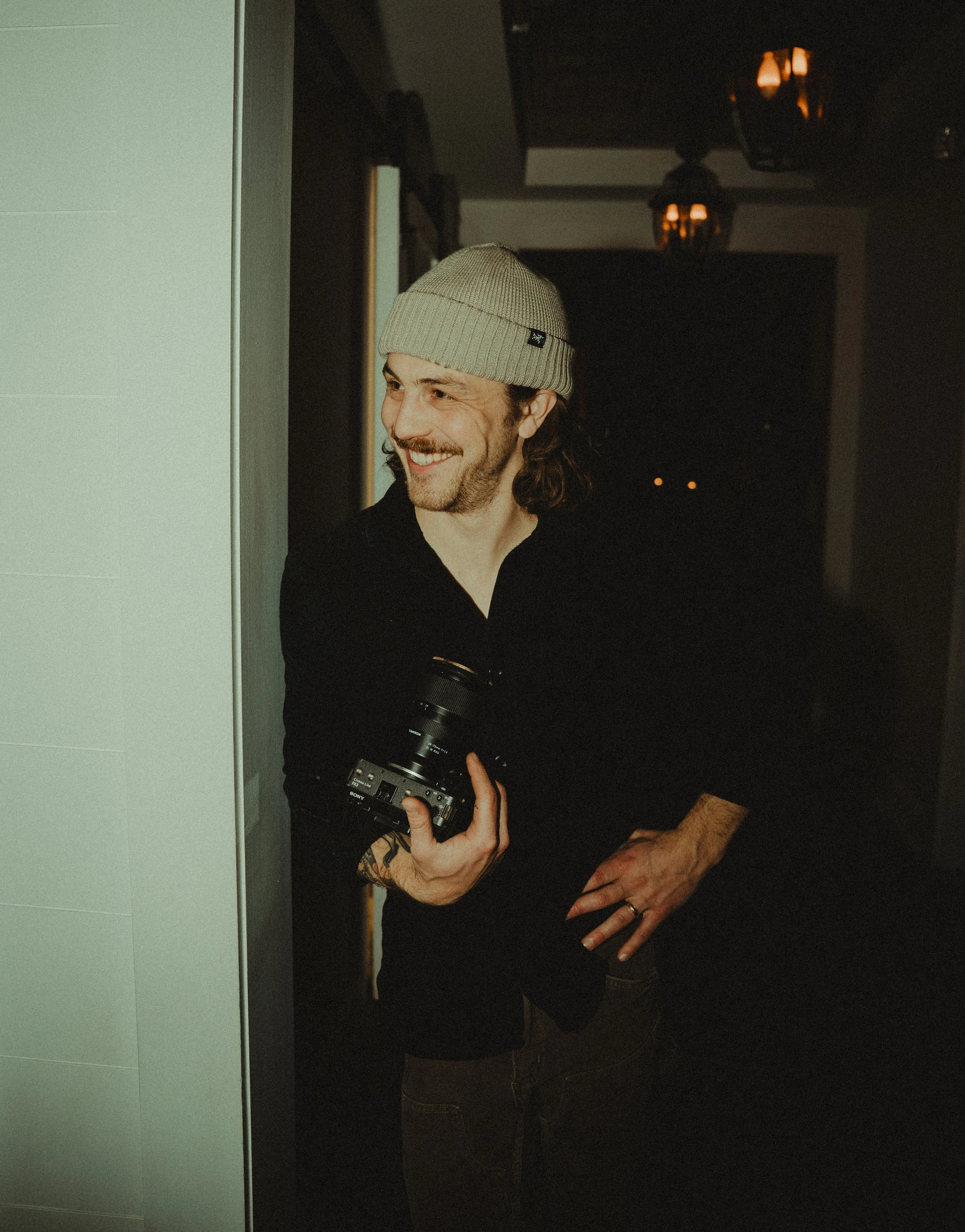 A smiling man with long hair, a beard, and a beanie hold a camera in his right hand. He's wearing a black shirt and has tattoos on his left arm, standing indoors near a wall with warm lighting in the background.