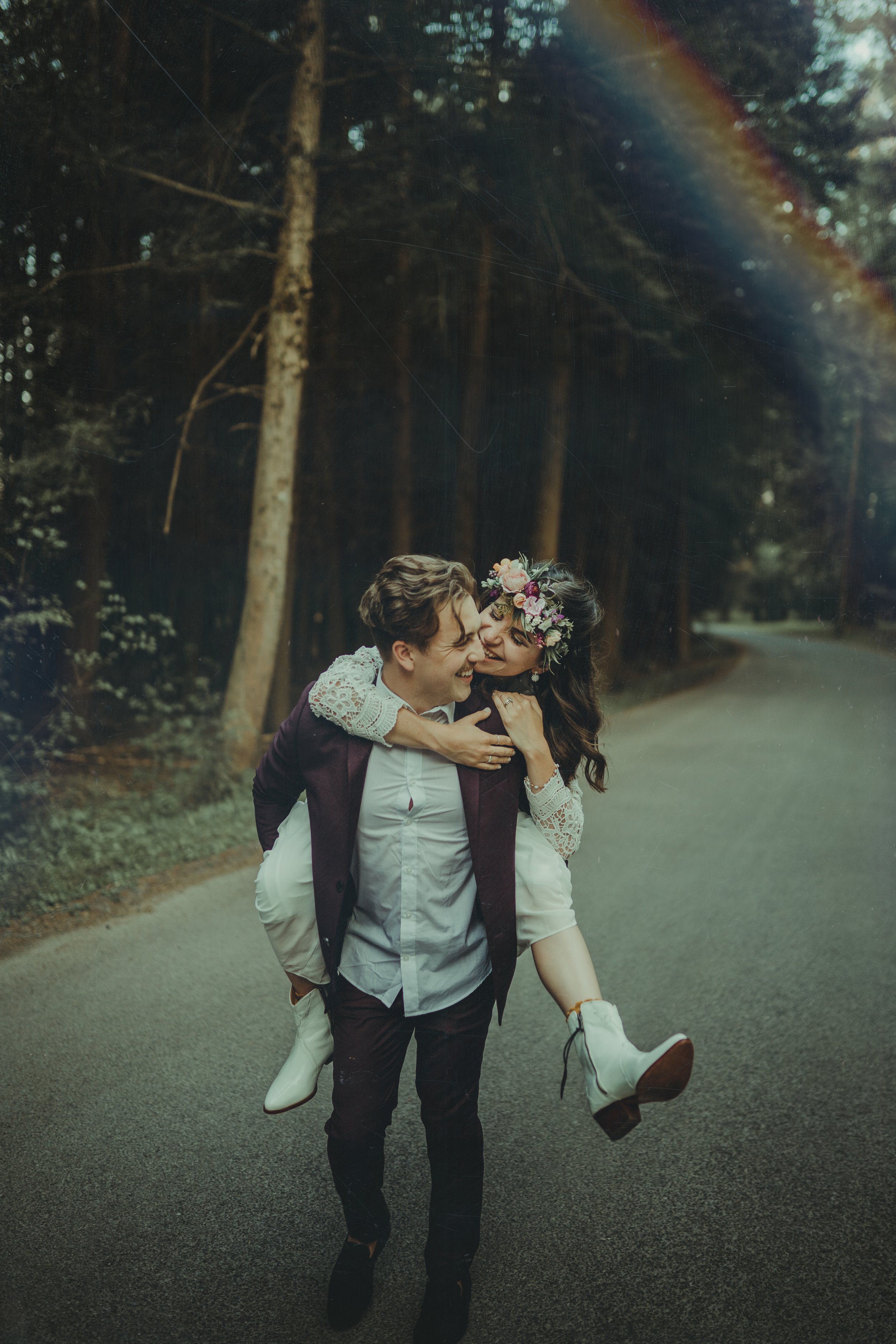 A joyful couple is outdoors on a scenic road surrounded by tall trees. The man is carrying the woman on his back, and they are both smiling and laughing. The woman is wearing a floral crown, a flowy white dress, and white boots, while the man is dressed in dark pants and a jacket. There's a rainbow visible in the sky.