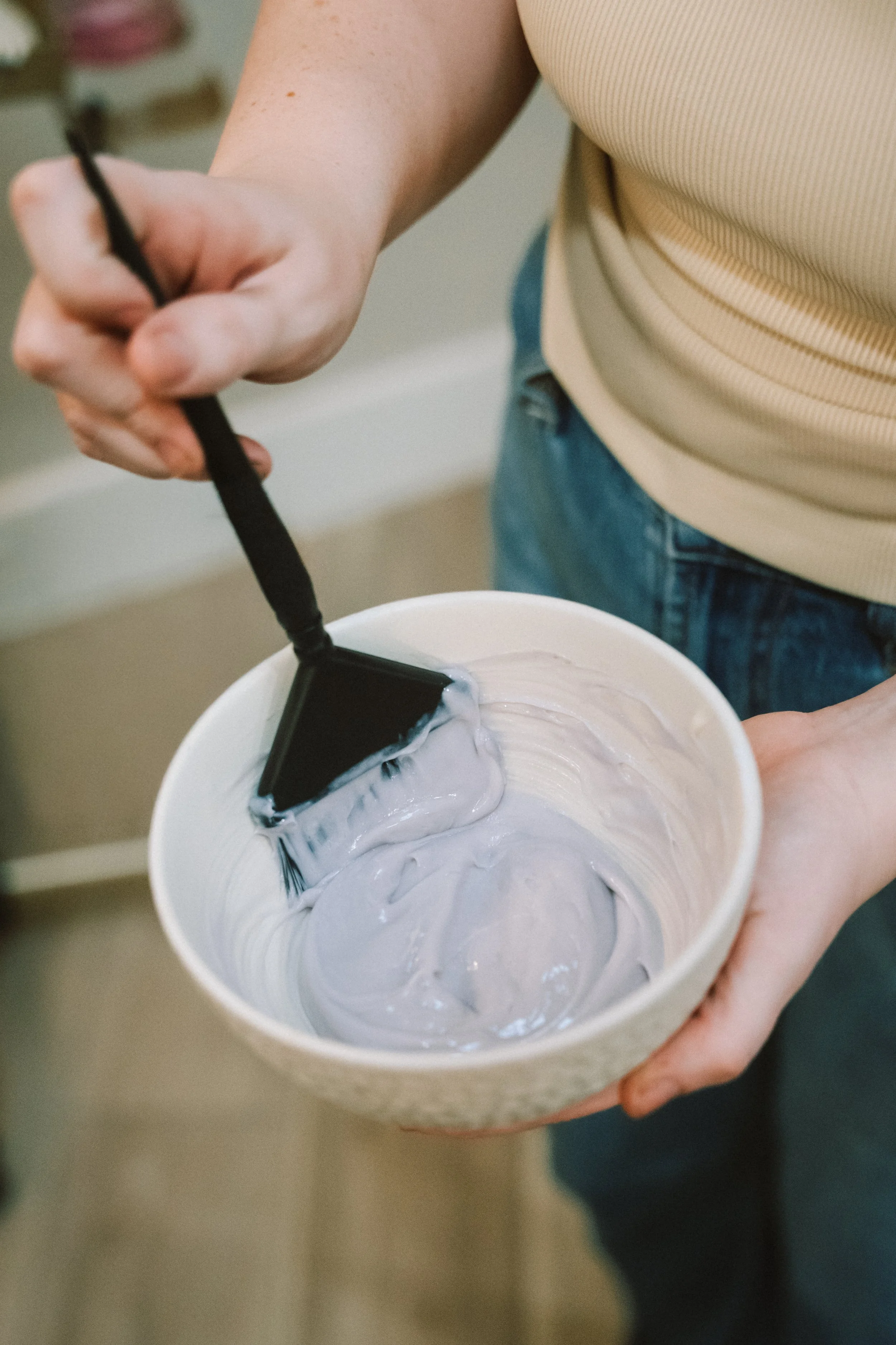 A person holding a white container with lavender-colored paint or slime inside, and a small brush or spatula stirring the mixture.