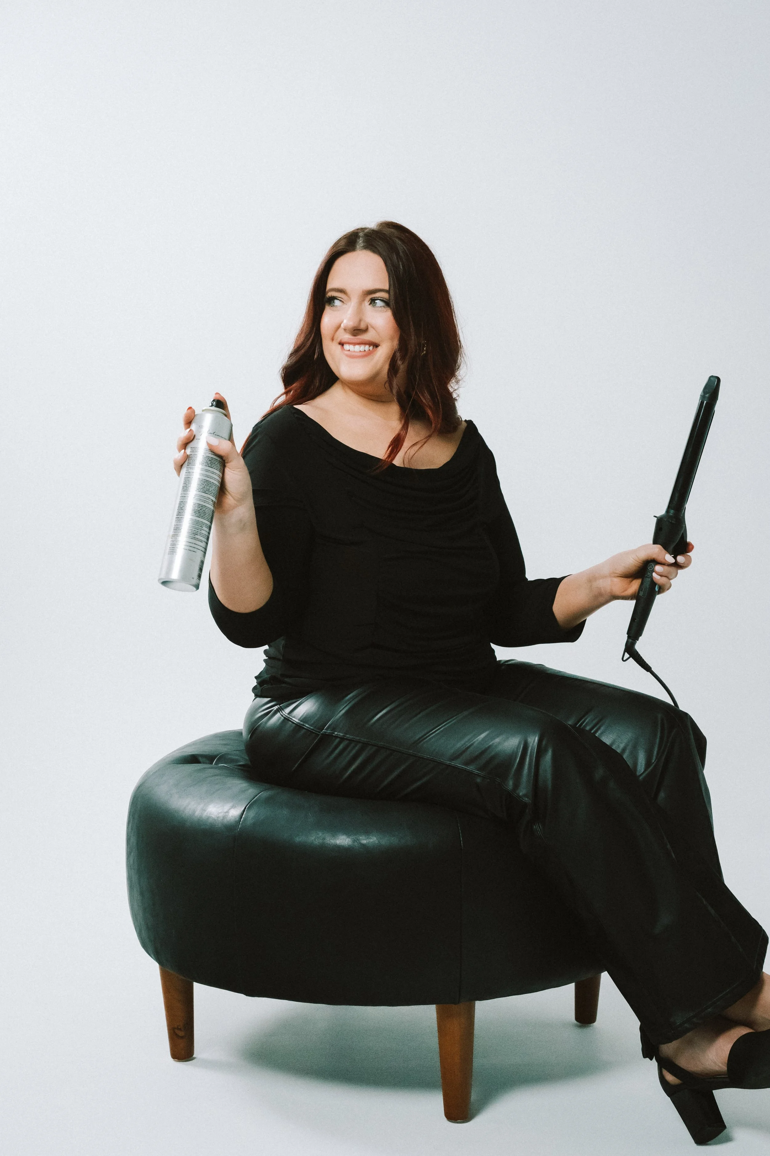 A woman sitting on a black leather ottoman, holding a hairspray can and a curling iron, smiling and looking to the side, against a plain white background.