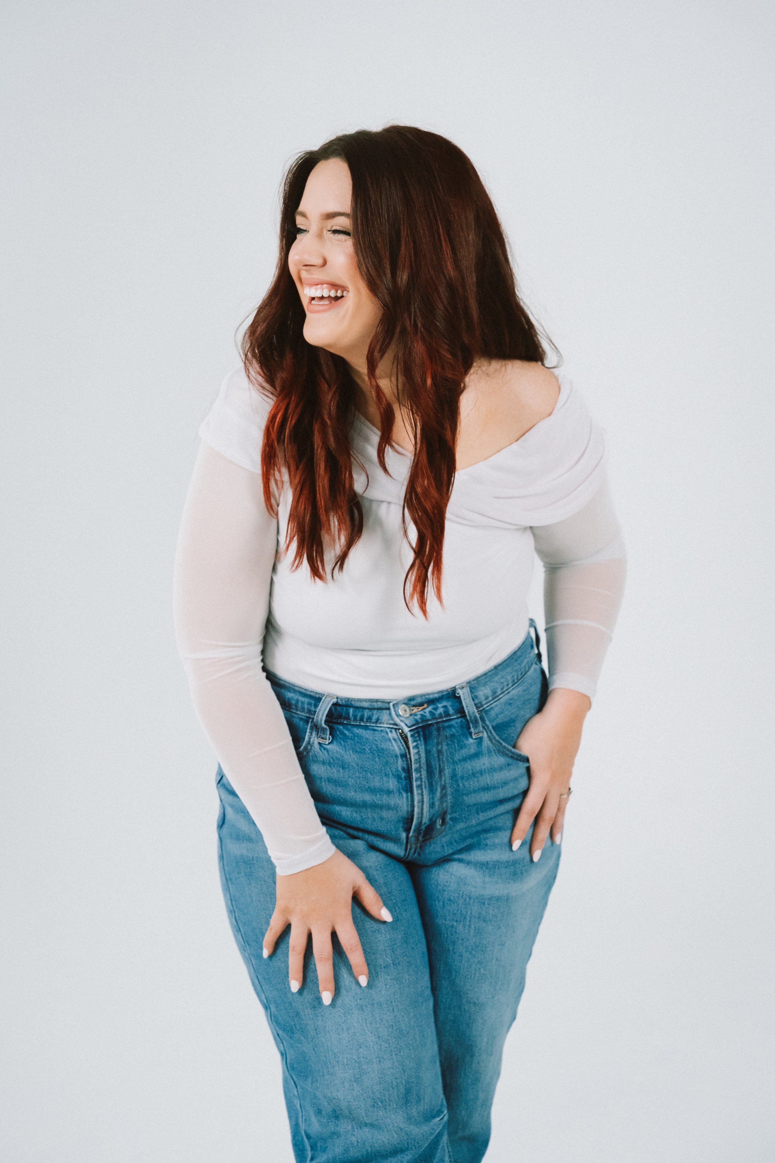 A woman with long, wavy brown hair with red highlights wearing a white off-the-shoulder top and blue jeans, laughing with her eyes closed against a plain white background.