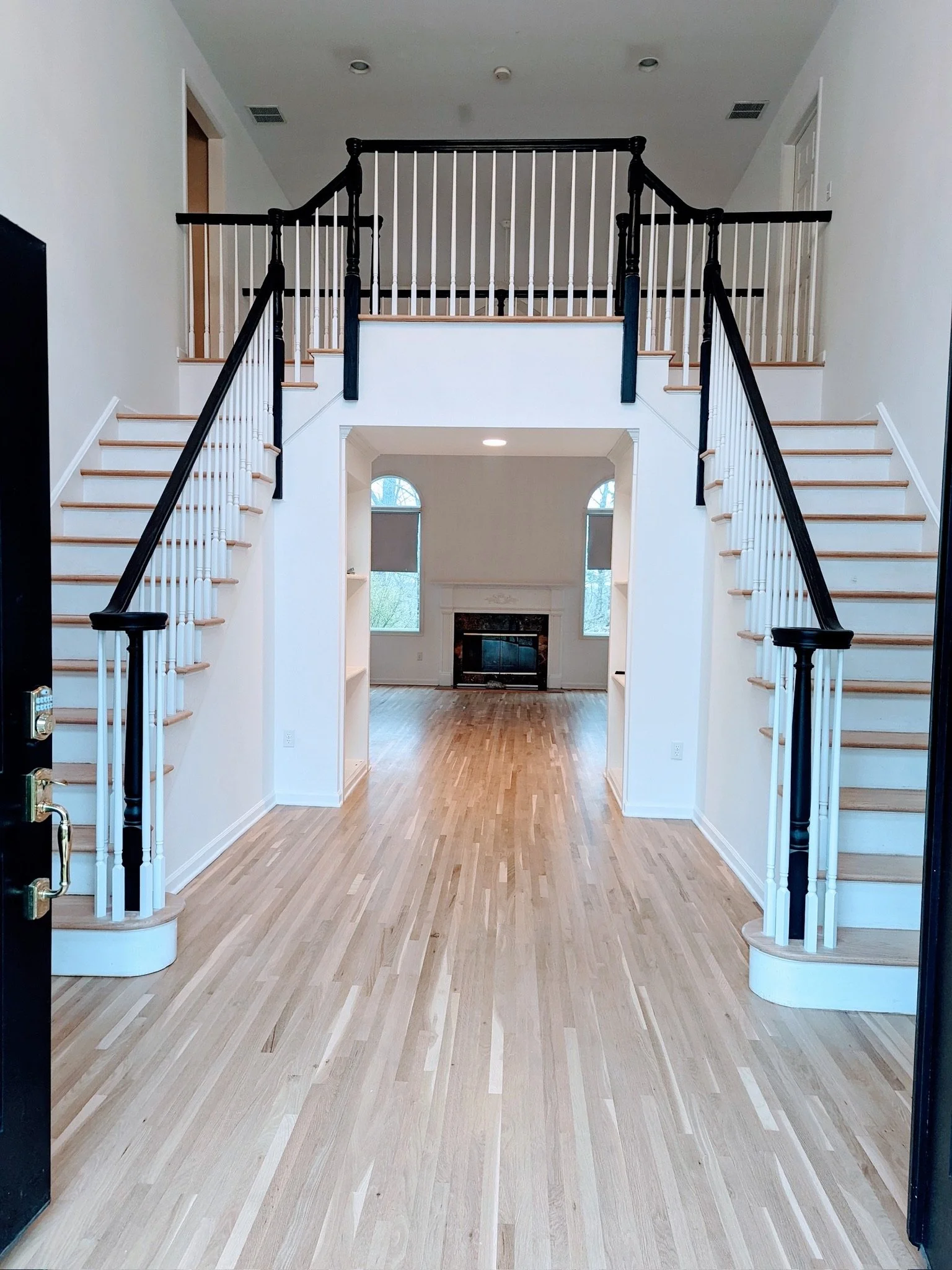 Two-story foyer with white walls, light wood floors, twin staircases with black railings, a fireplace, and a large arched window.