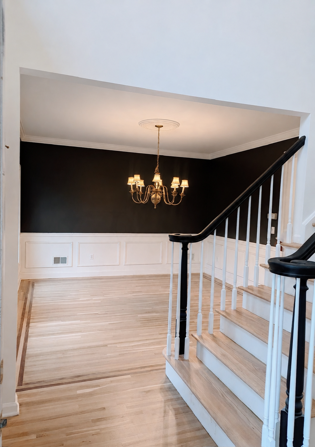 Dining area with a dark accent wall, white wainscoting, brass chandelier, hardwood floors, and black-and-white staircase railings.