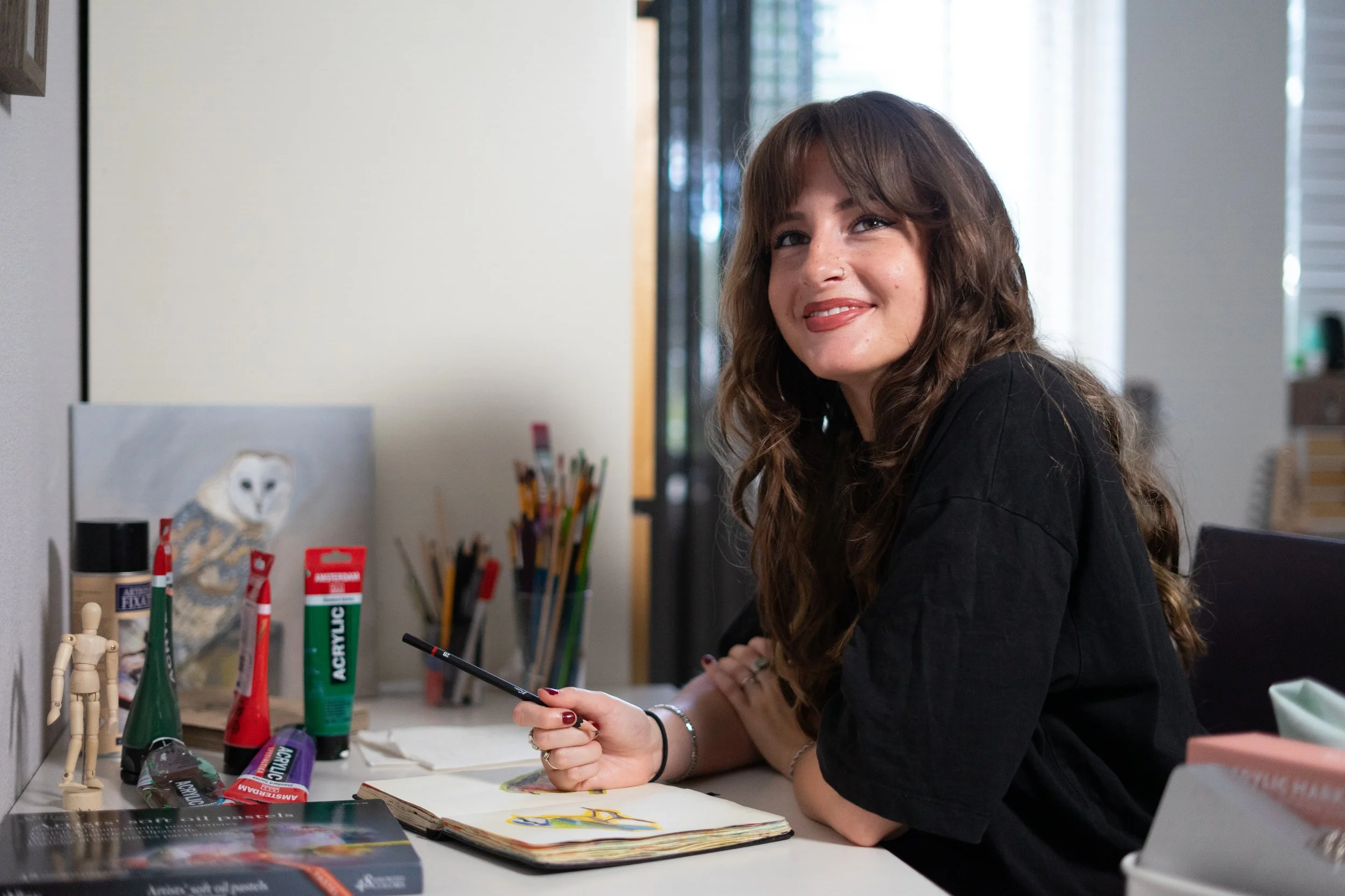 A woman with long, wavy brown hair and a nose piercing sitting at a desk, smiling, surrounded by art supplies and drawing materials.