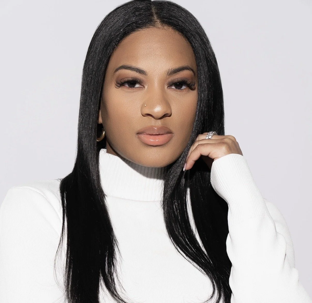 A woman with long black hair, wearing a white turtleneck sweater, looking directly at the camera with a neutral expression.