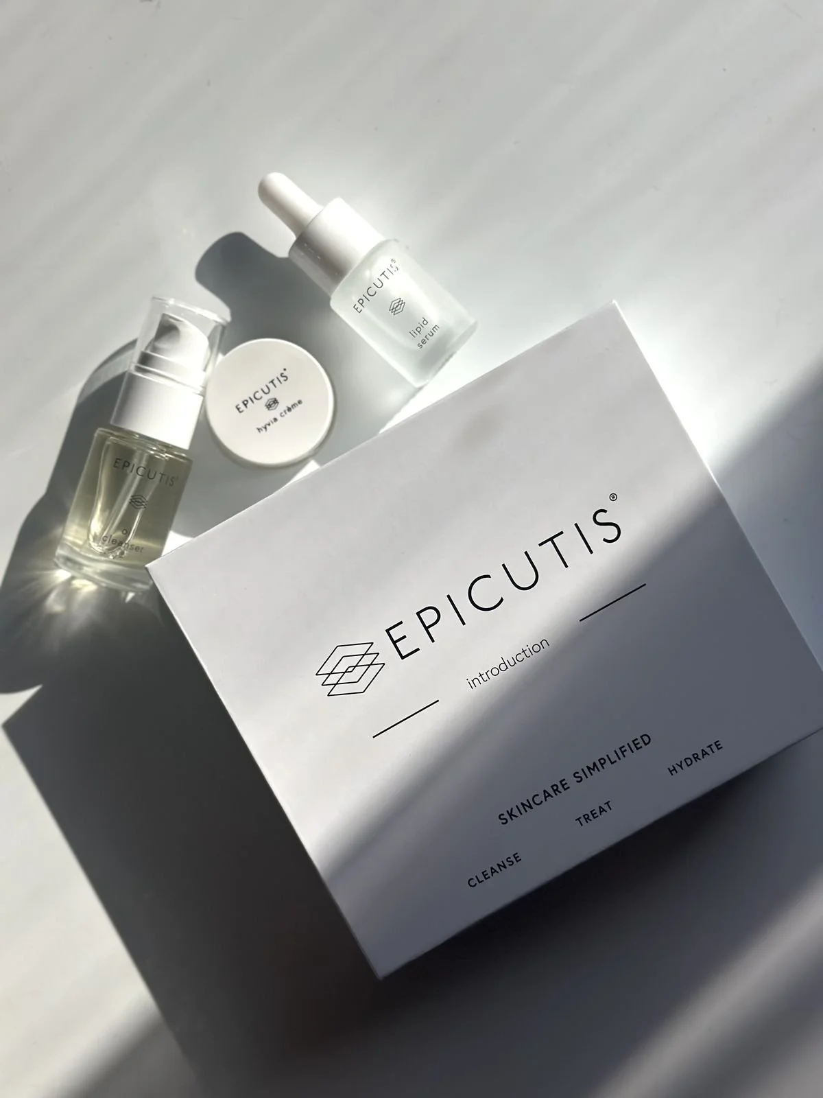 Skincare products and a box from the brand Epicutis, including a cleanser, a lipid serum, and a nourishing cream, all arranged on a white surface with shadows from natural light.