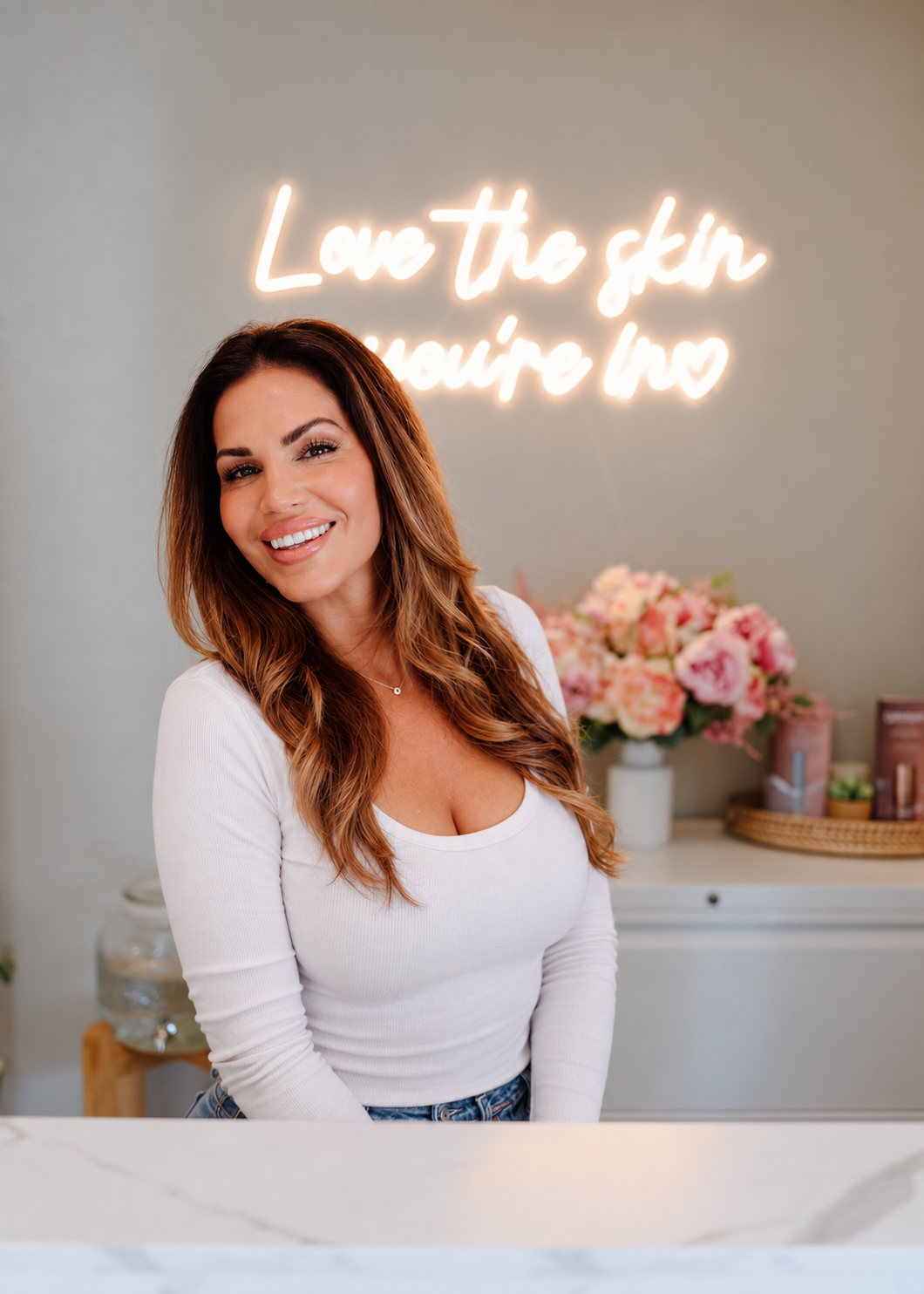Santa Cruz esthetician Chrissy Jacobs smiling in front of her “Love the skin you’re in” neon sign
