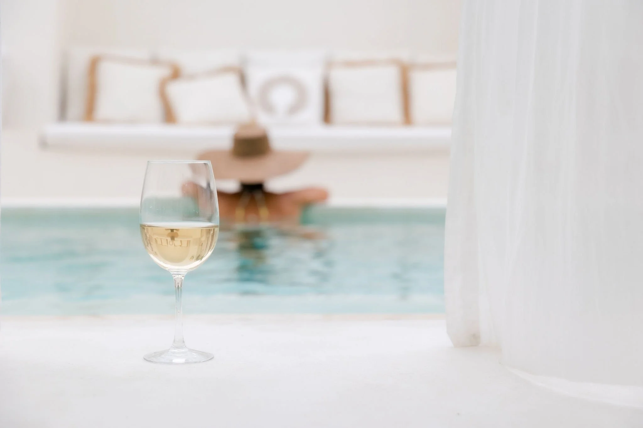 A glass of white wine on a white surface, with a blurred background showing a white room with chairs and a table.