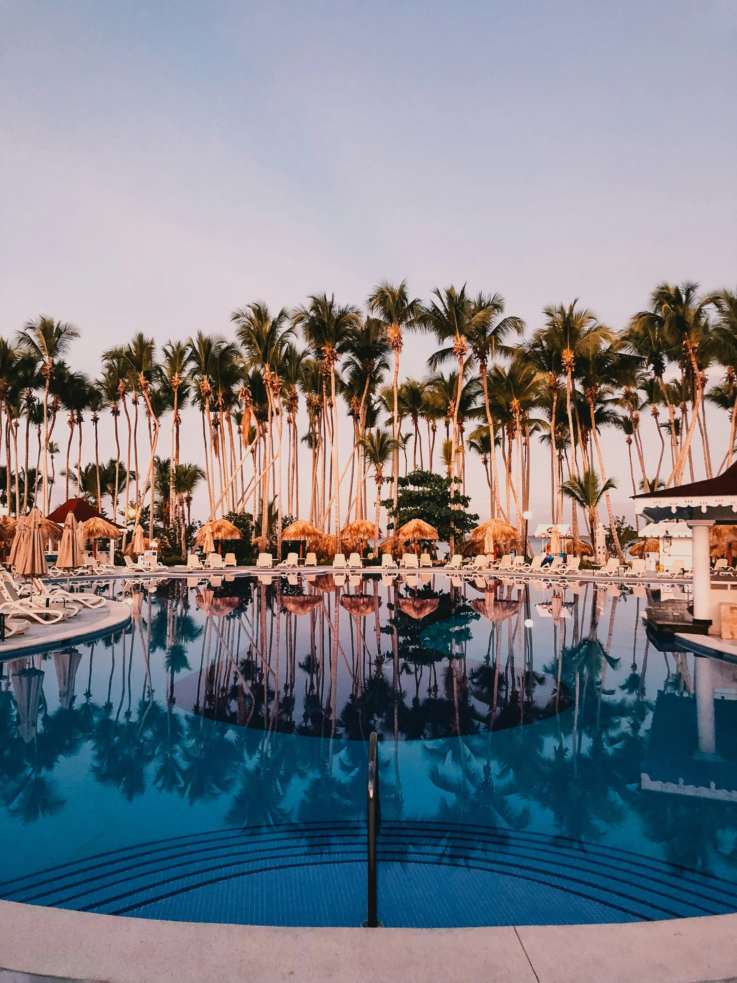 A swimming pool with a curved edge reflecting surrounding palm trees and sky during sunset, with poolside chairs and straw umbrellas.