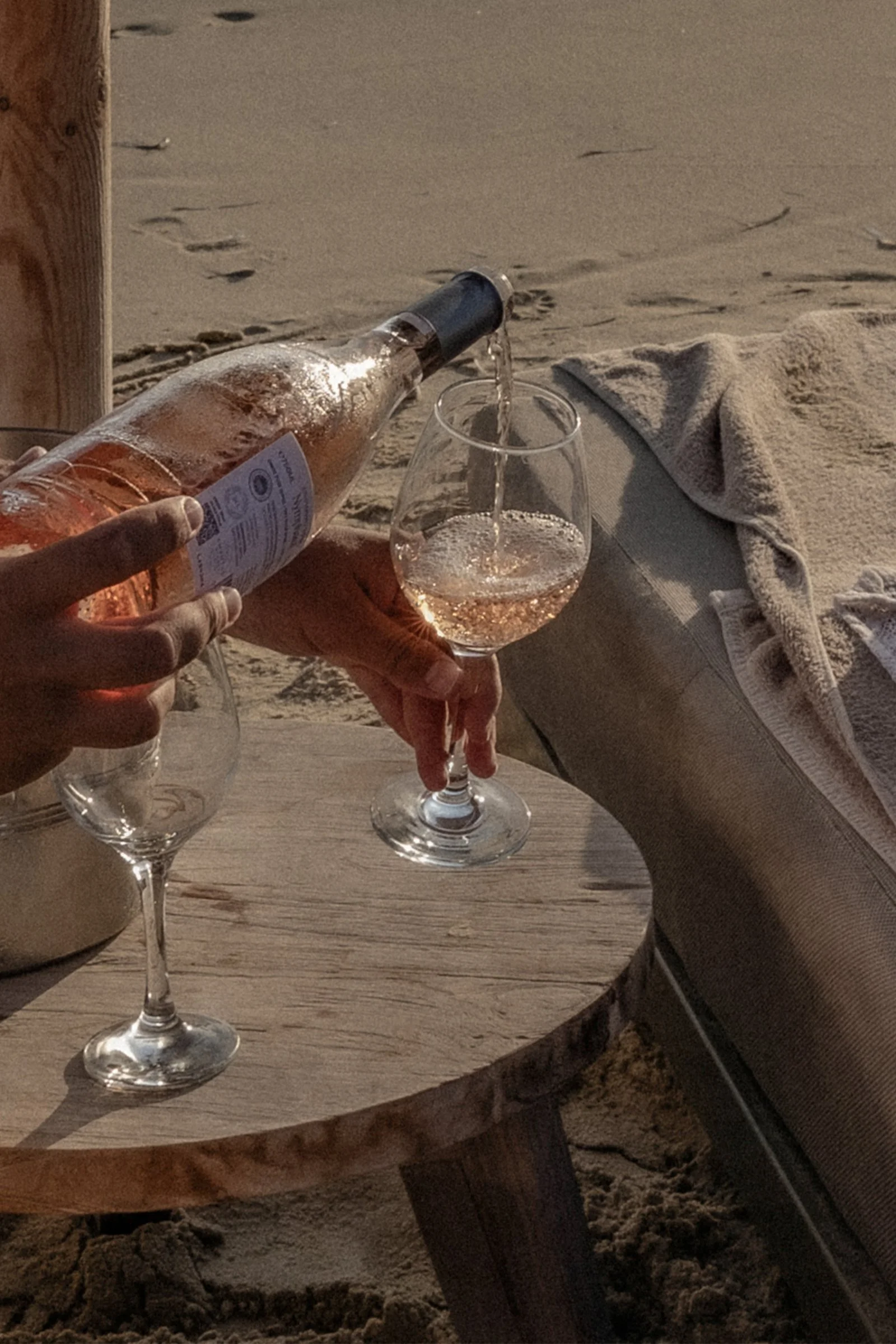 A person pouring a glass of rosé wine on a beach, with sand, a wooden table, and a gray towel nearby.
