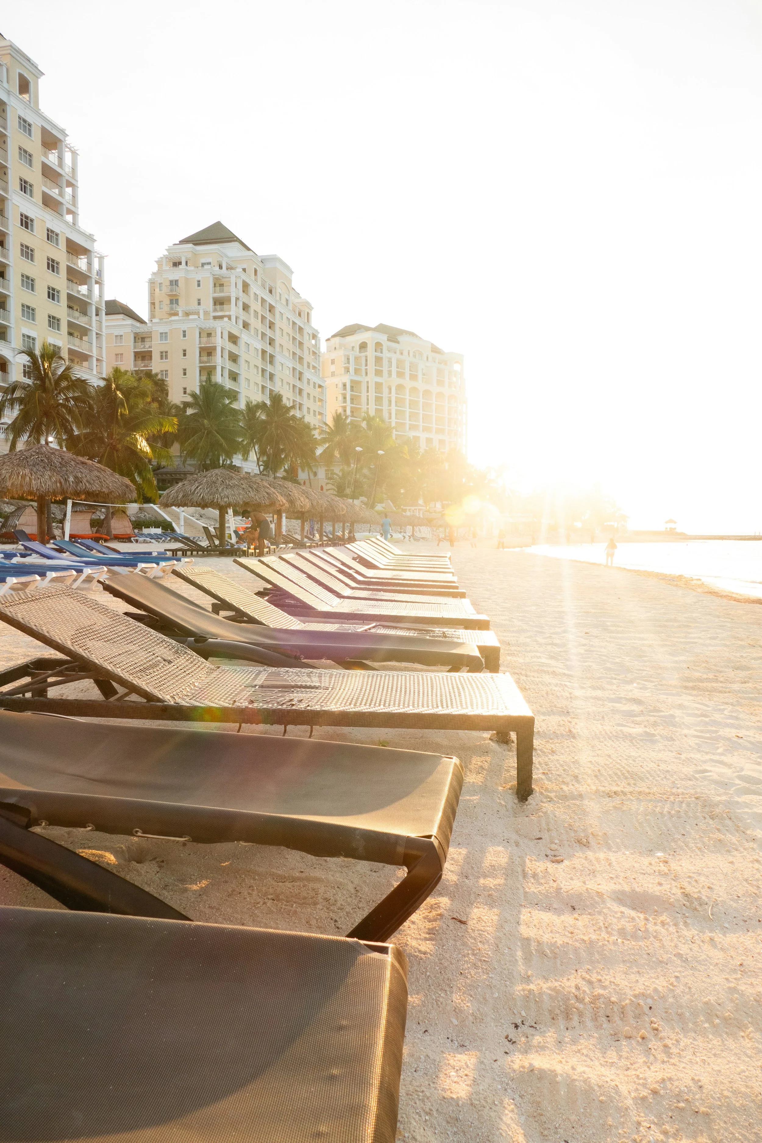 Beach scene with lounge chairs and umbrellas, tall buildings in the background, palm trees, and the sun setting over the ocean.