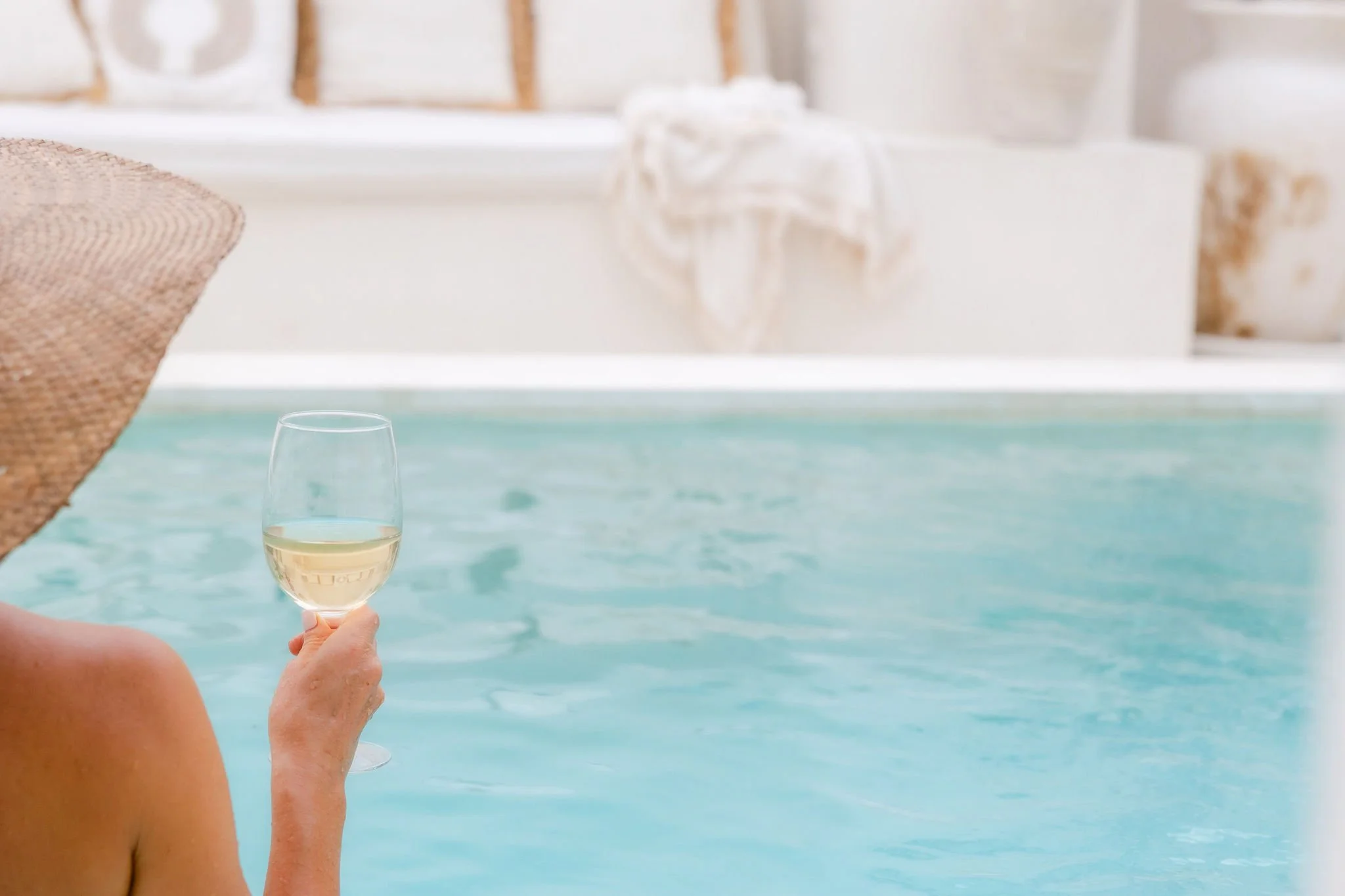 Person by a pool holding a glass of white wine with a seating area in the background.