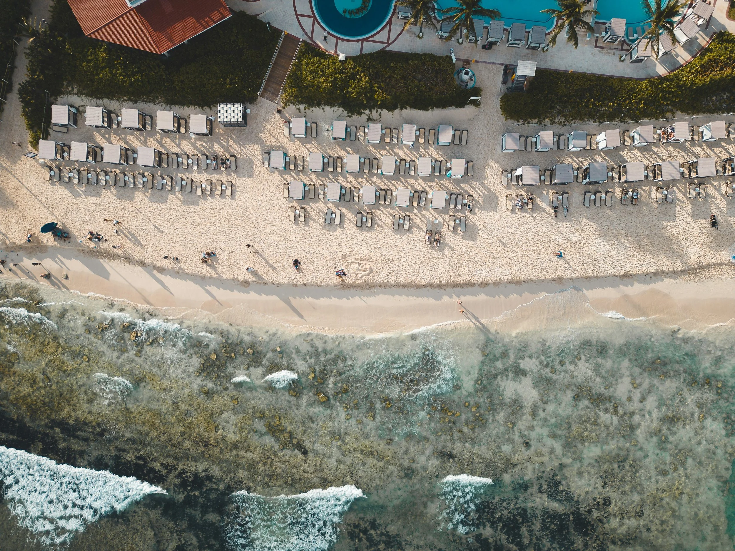 Aerial view of a beach with lounge chairs, umbrellas, a swimming pool, palm trees, and people walking along the shore and on the sidewalk.