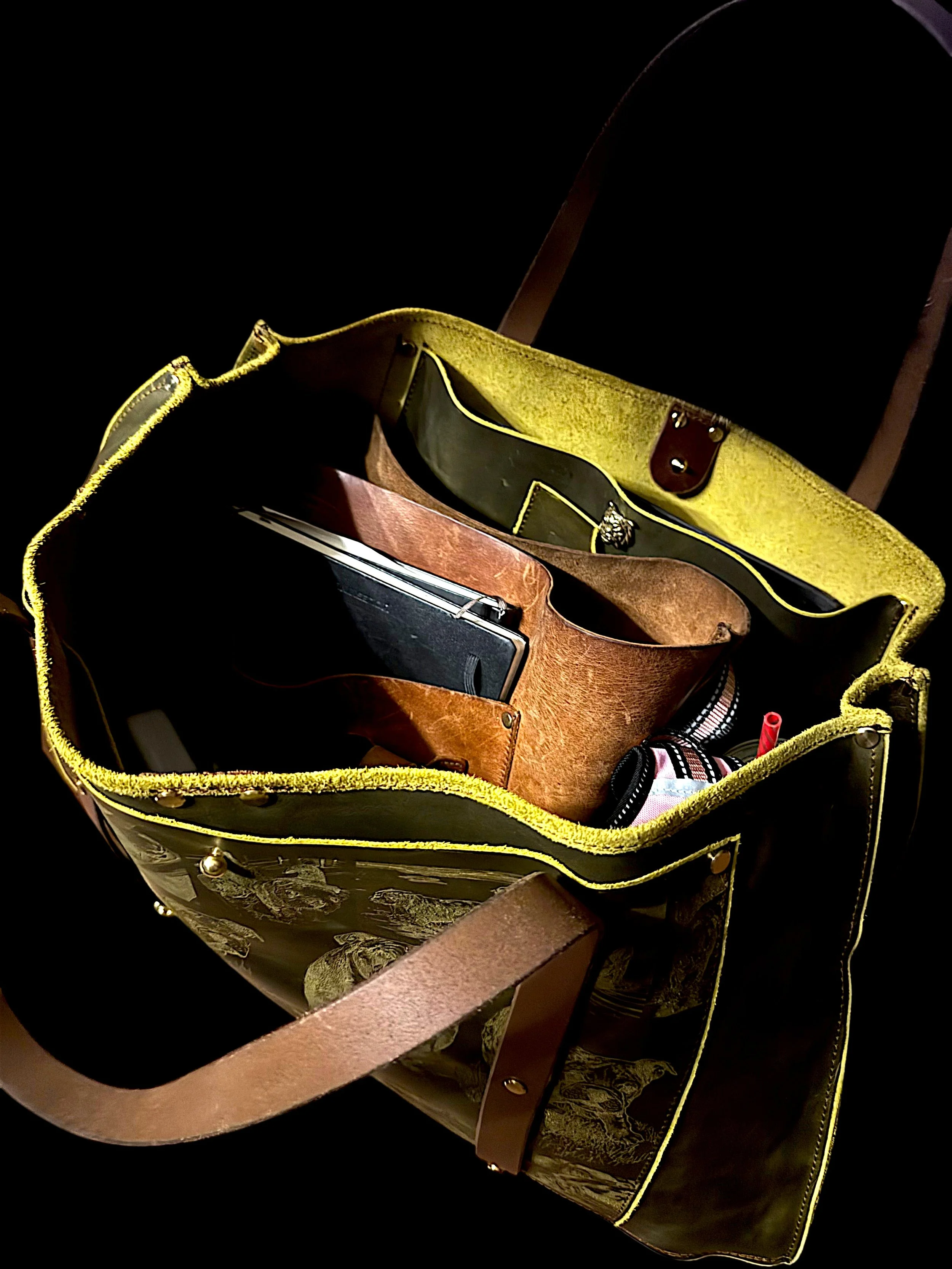 Open leather bag with yellow interior, containing items such as a notebook, pen, and a small pouch, placed on a black background.