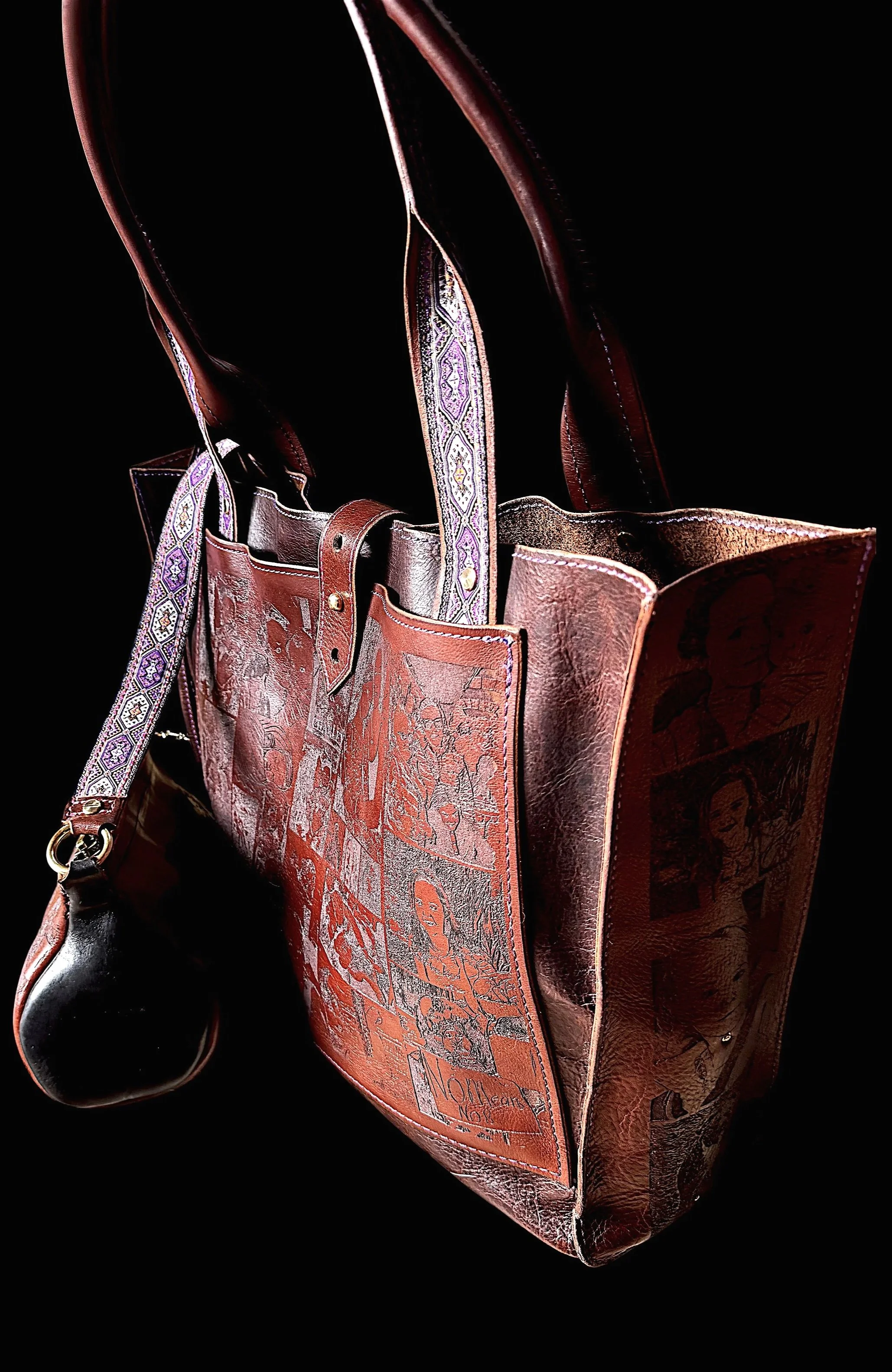 A brown leather tote bag with graphic art and colorful details, featuring a detachable patterned strap, positioned against a black background.