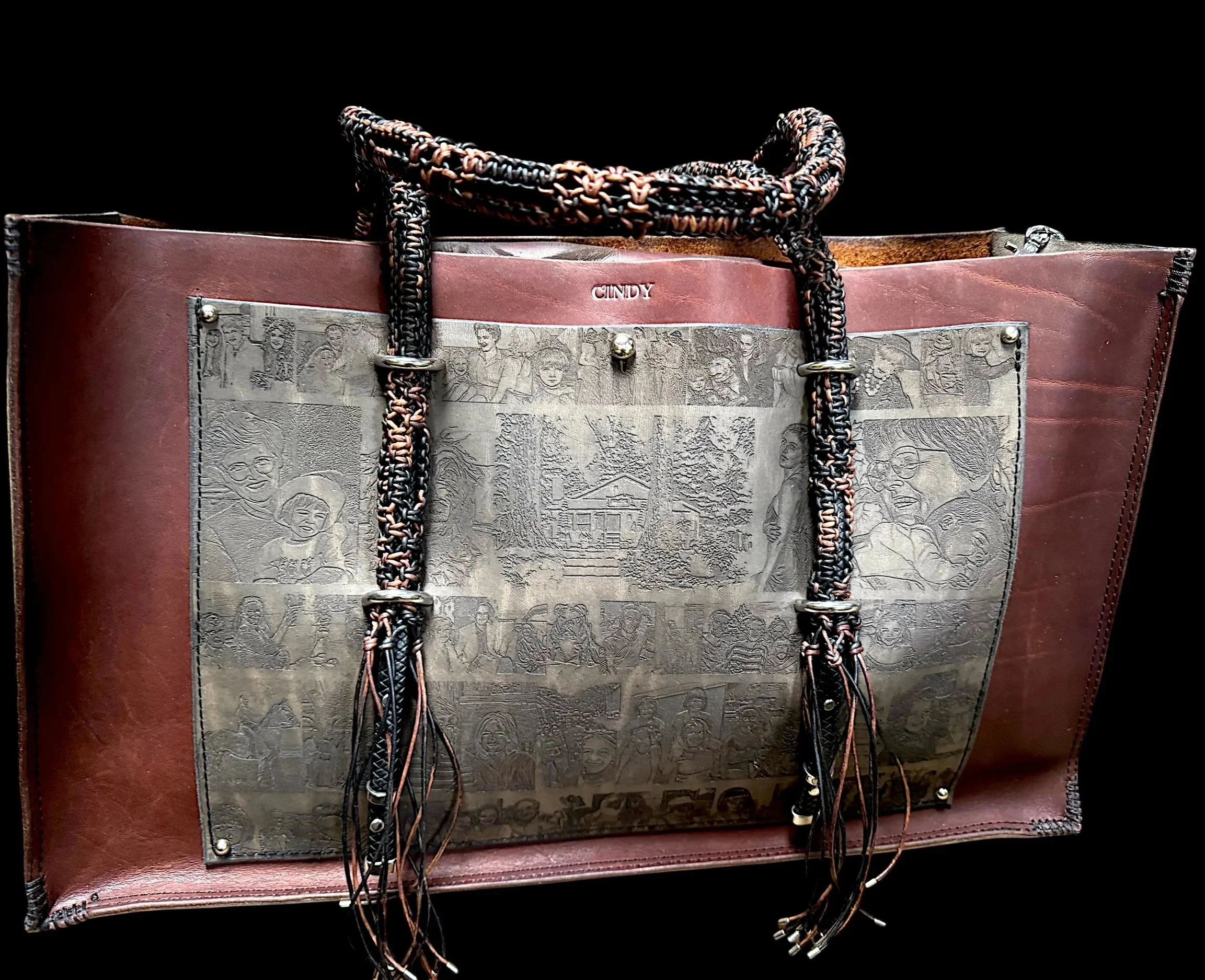 Close-up of a brown leather tote bag with a background of black and gray drawn images of children and adults on the front pocket. The bag has braided handles with black, pink, and beige strands. The word 'CINDY' is embossed above the pocket.