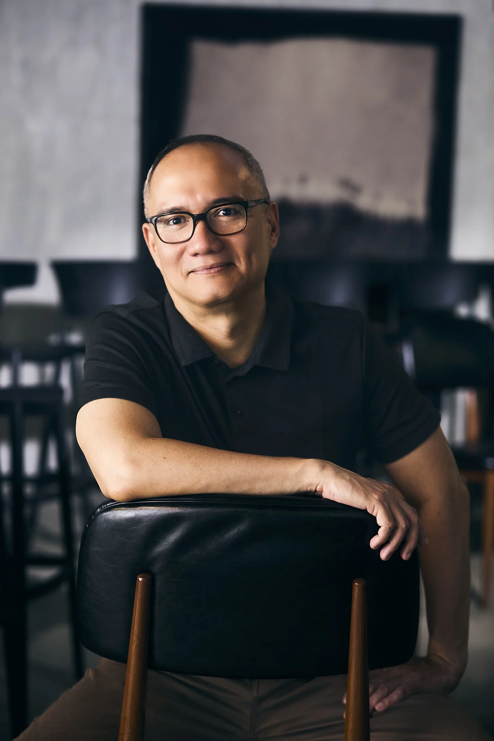 A man with glasses smiling at the camera, sitting in a room with a gray wall, an abstract painting in the background, and surrounded by black chairs.