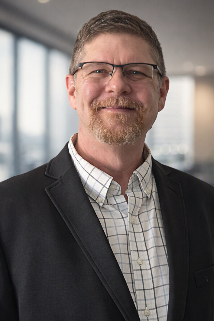 A middle-aged man with glasses, a beard, and a mustache, wearing a black blazer and a white checked shirt, smiling in an office setting.