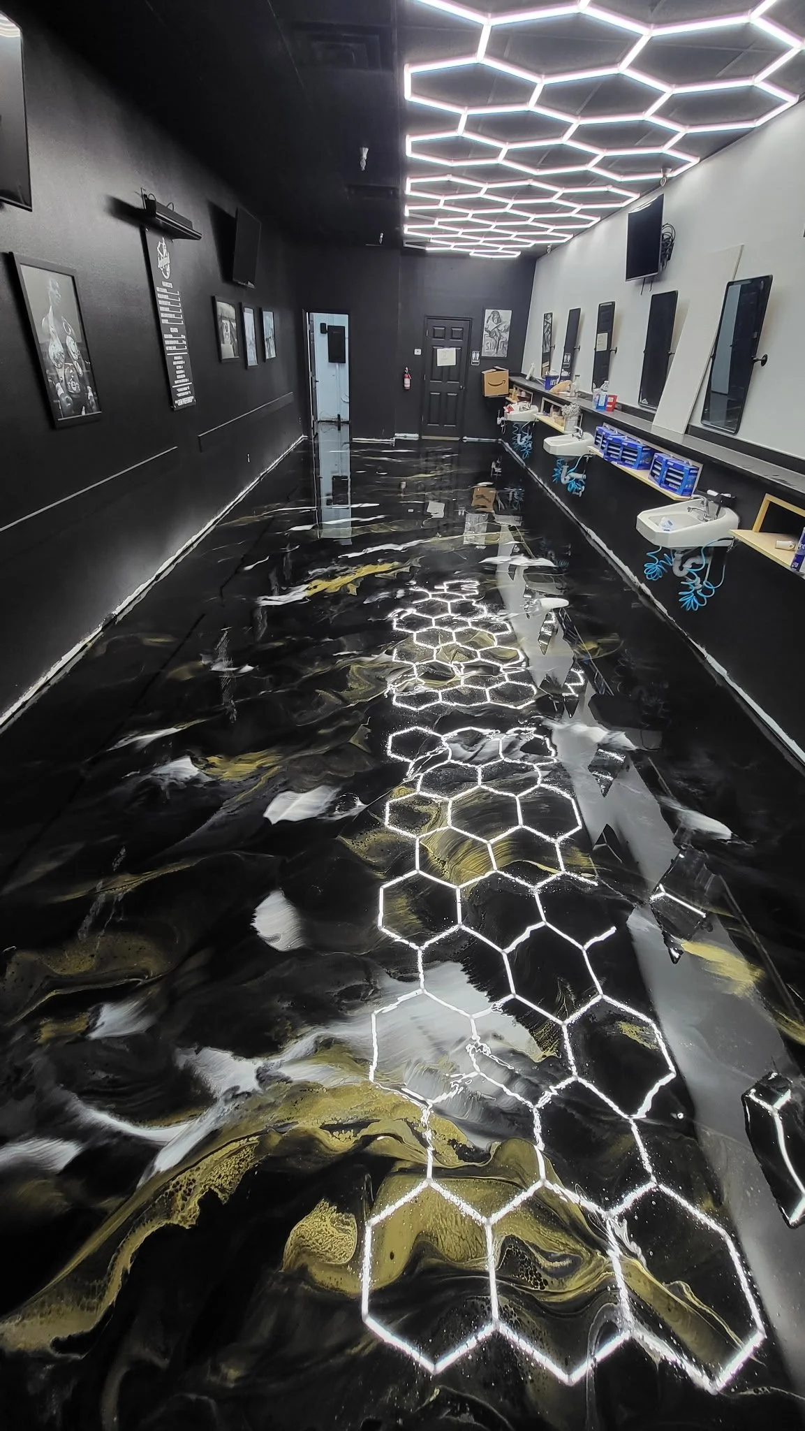 Interior of a modern barbershop or salon with black and white walls, hexagon-shaped LED ceiling lights, and a shiny black marble floor with white and gold veining, reflecting the ceiling lights.