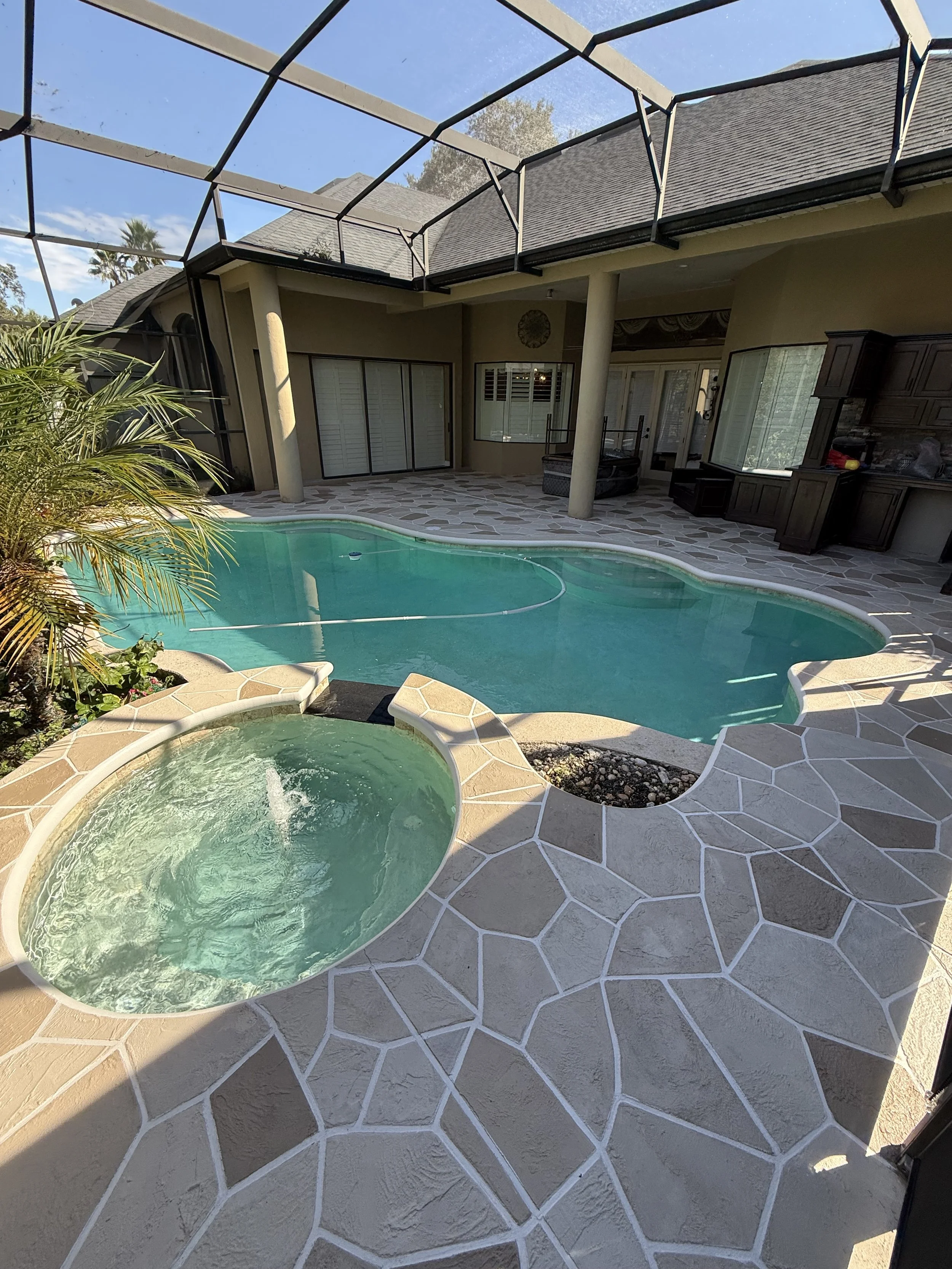 Backyard with a swimming pool, hot tub, and a covered patio with sliding glass doors, surrounded by a fence and green plants, with a clear blue sky above.