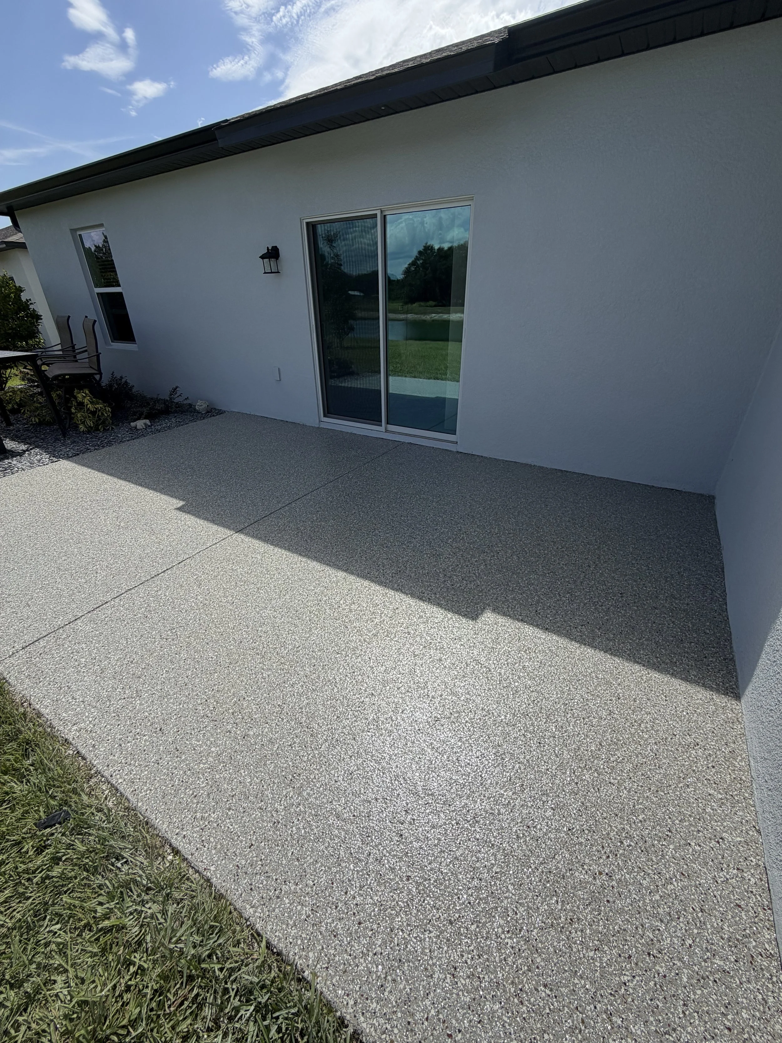 A backyard patio with a textured concrete surface, a white house wall with a window and sliding door, outdoor seating to the side, and a grassy area with trees reflected in the door's glass, under a partly cloudy sky.