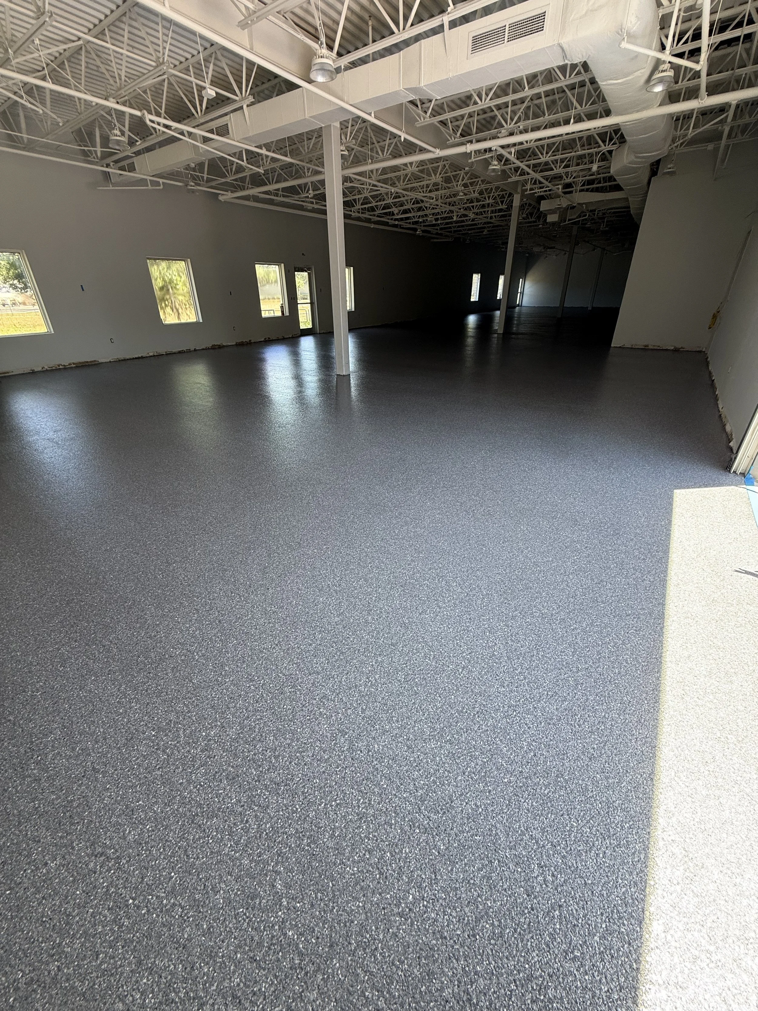 Interior of a commercial space under construction with gray speckled epoxy flooring, white walls, and an exposed ceiling with metal beams, ductwork, and lighting fixtures.