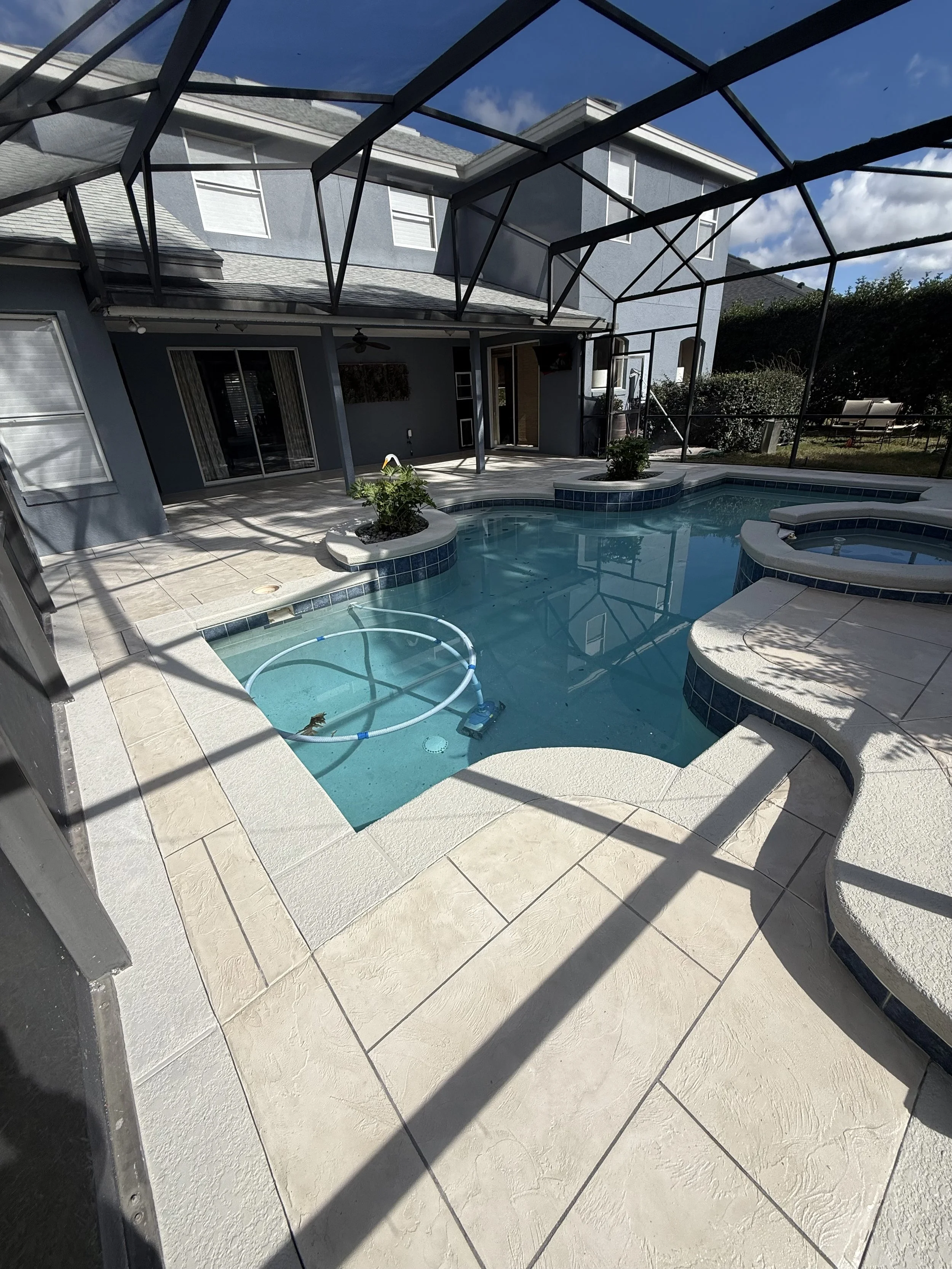 Backyard with swimming pool, screened enclosure, patio area, house with closed blinds and sliding glass door, outdoor seating, and some bushes and trees in the background.
