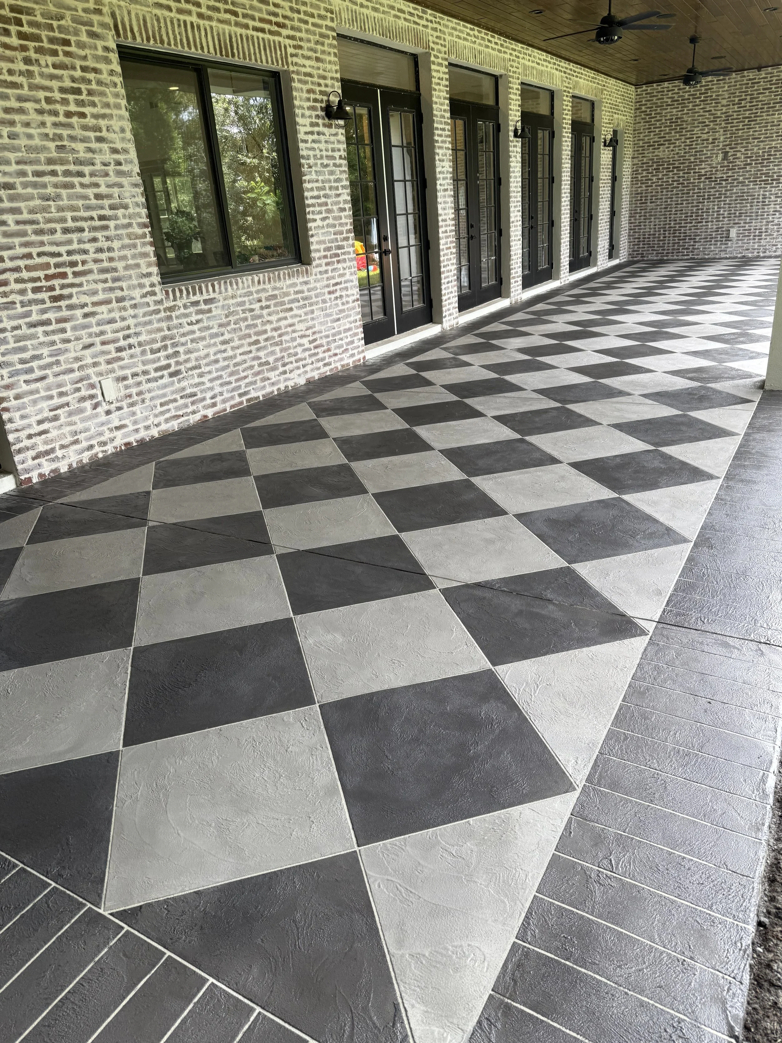 Covered porch area with checkerboard-patterned black and white tile flooring, brick walls, and black-framed glass doors and windows. There are three black wall-mounted lamps and ceiling fans on a wooden ceiling.